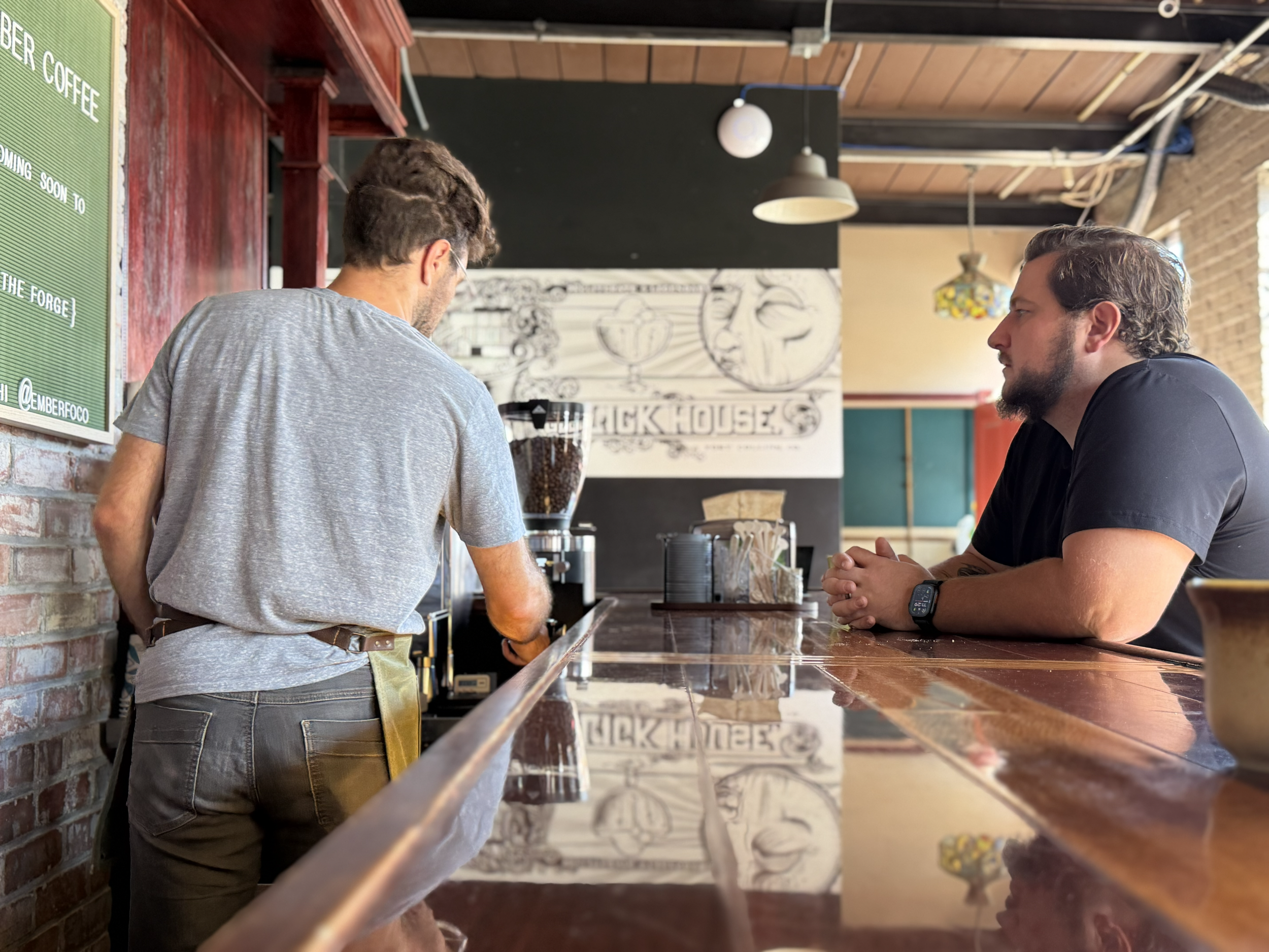 Two men sitting and talking at a wood bar counter inside a coffee shop, with a coffee grinder and a sign board in background.