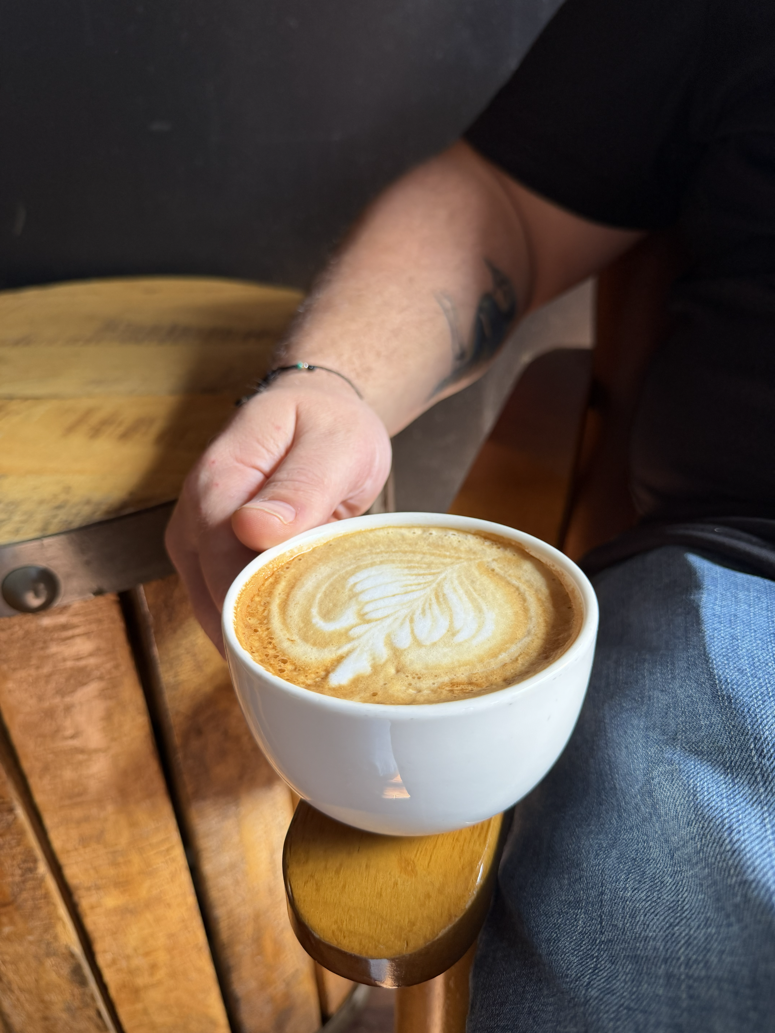 A person holding a white mug of coffee with latte art, sitting on a wooden bench wearing dark clothing and jeans.