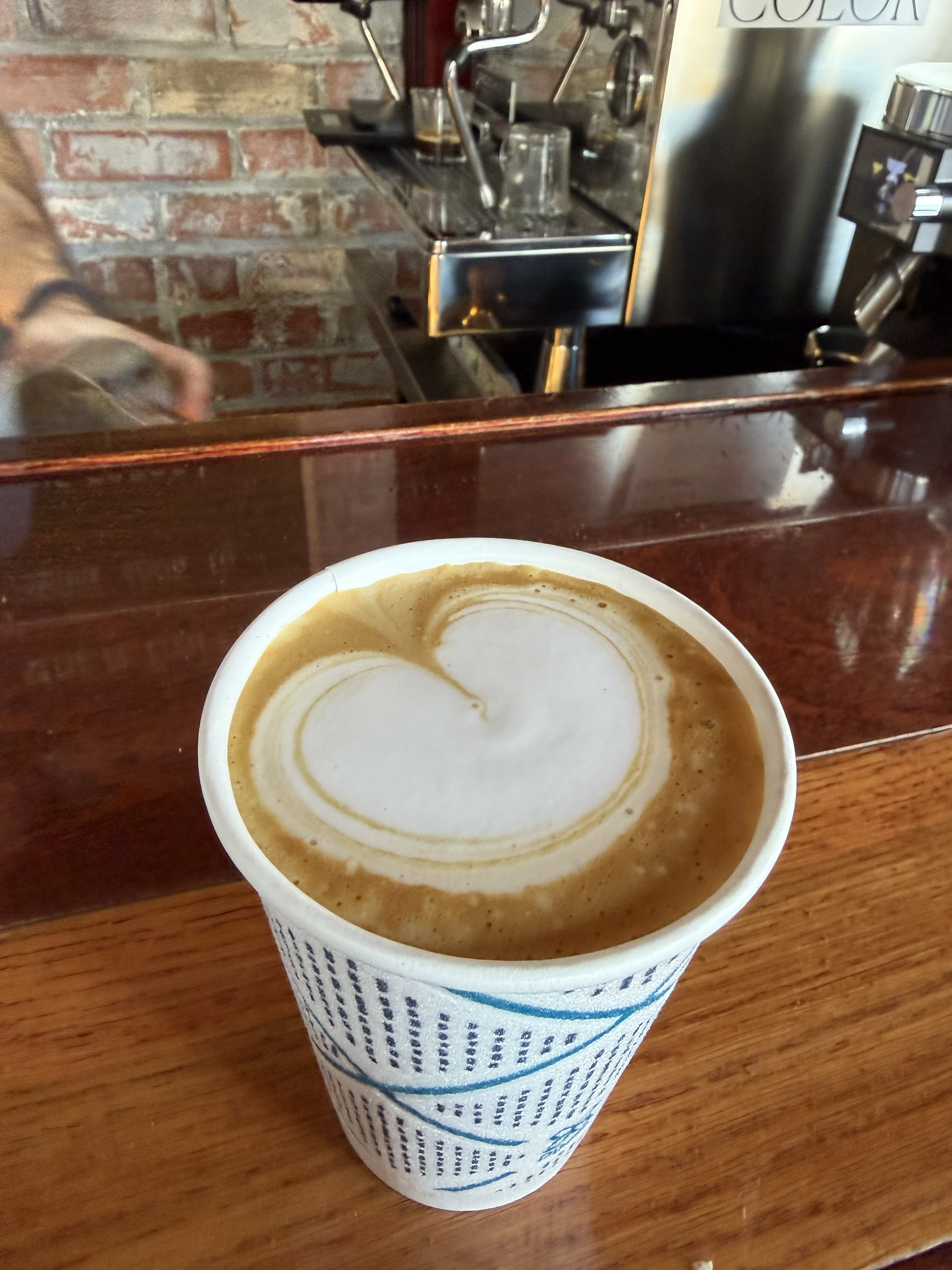 Close-up of a disposable coffee cup with latte art on top of foam, placed on a wooden table in a coffee shop, with a brick wall and coffee brewing equipment in the background.