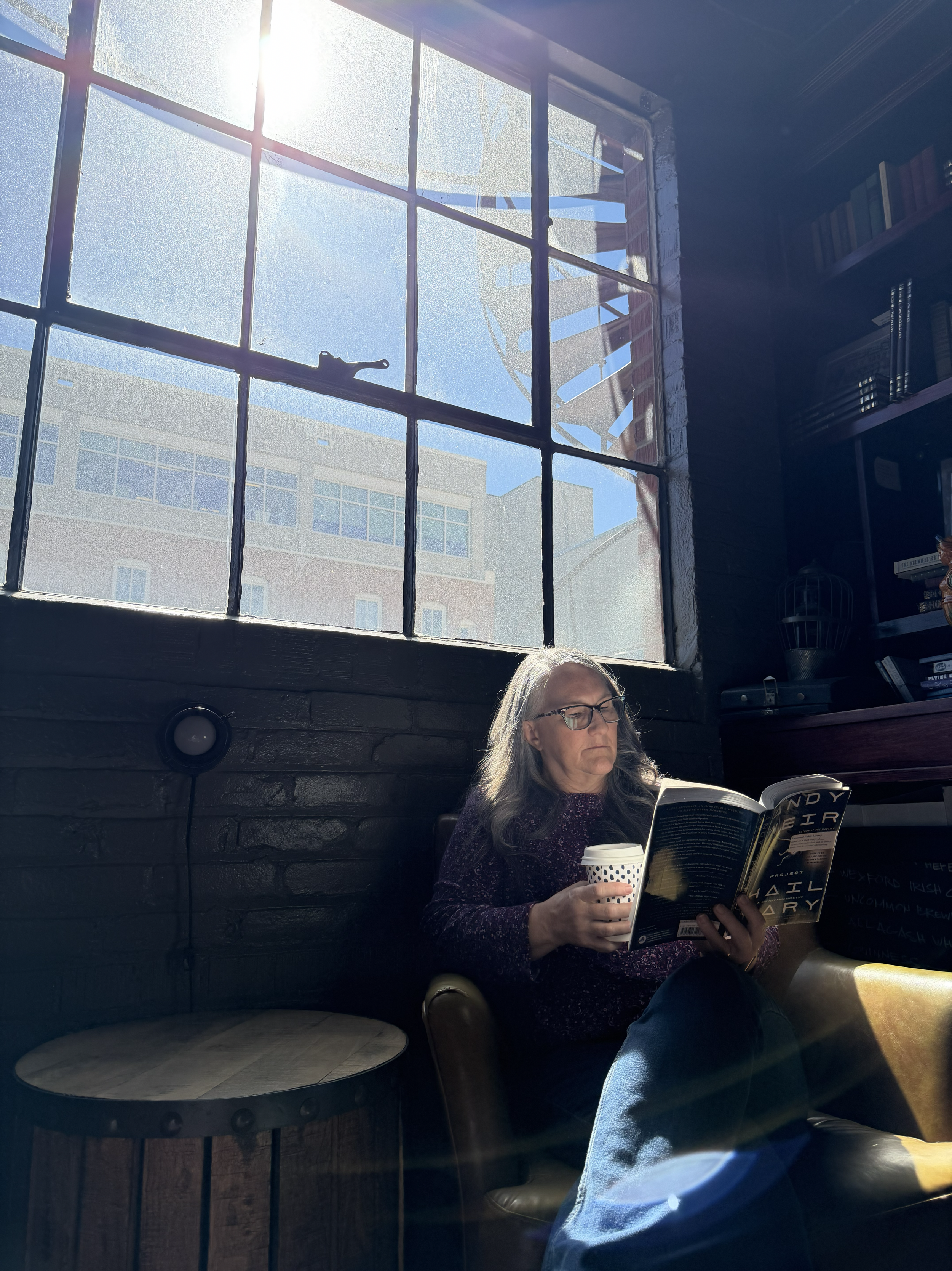 A woman sitting next to a window in a cozy indoor space, reading a magazine and holding a cup of coffee, with sunlight streaming through the window.