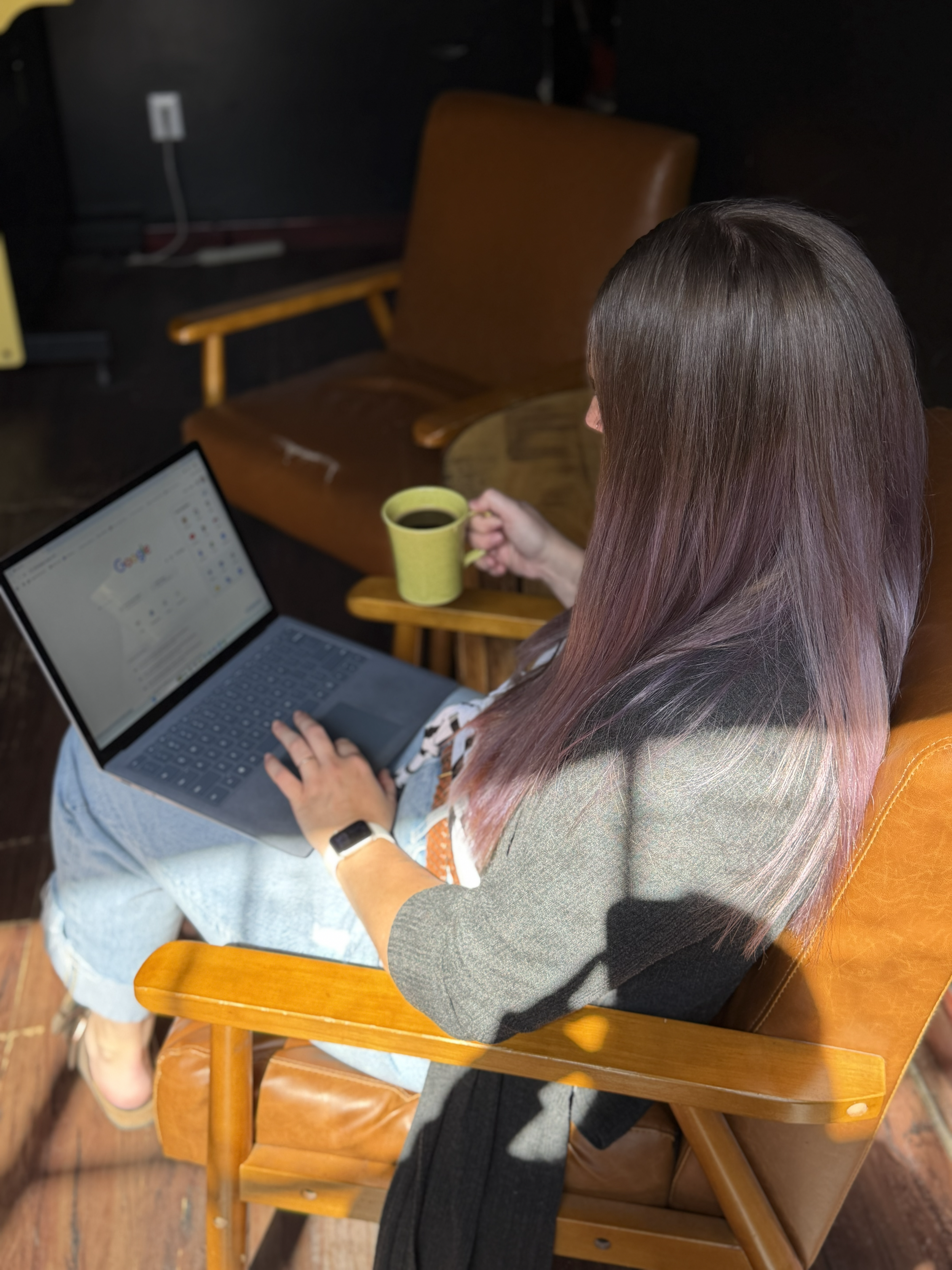 A woman with long, brown hair sitting on a wooden chair, holding a yellow mug of coffee, working on a laptop.