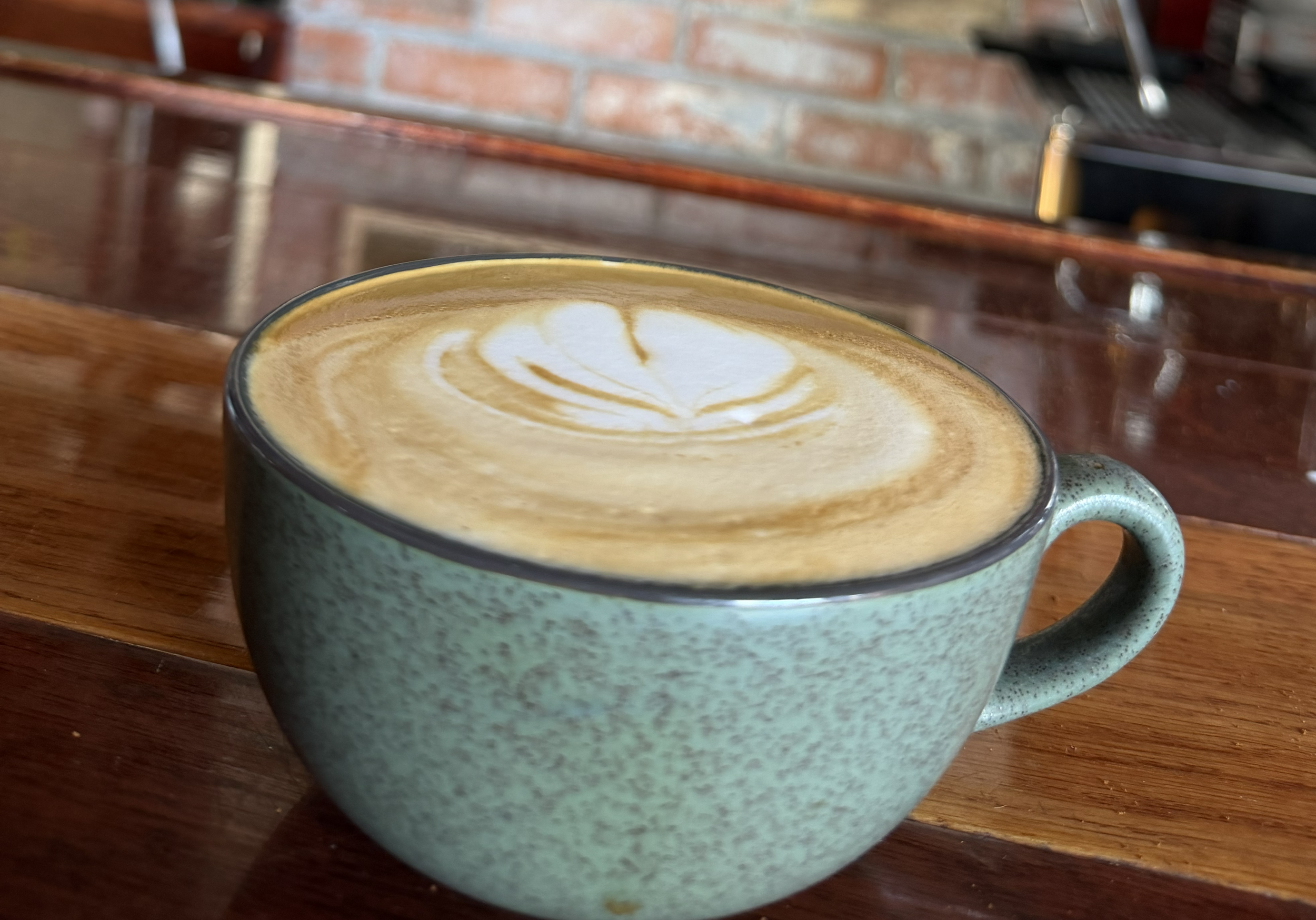 A ceramic cup filled with a latte showcasing latte art on a wooden table, with a brick wall and kitchen utensils in the background.