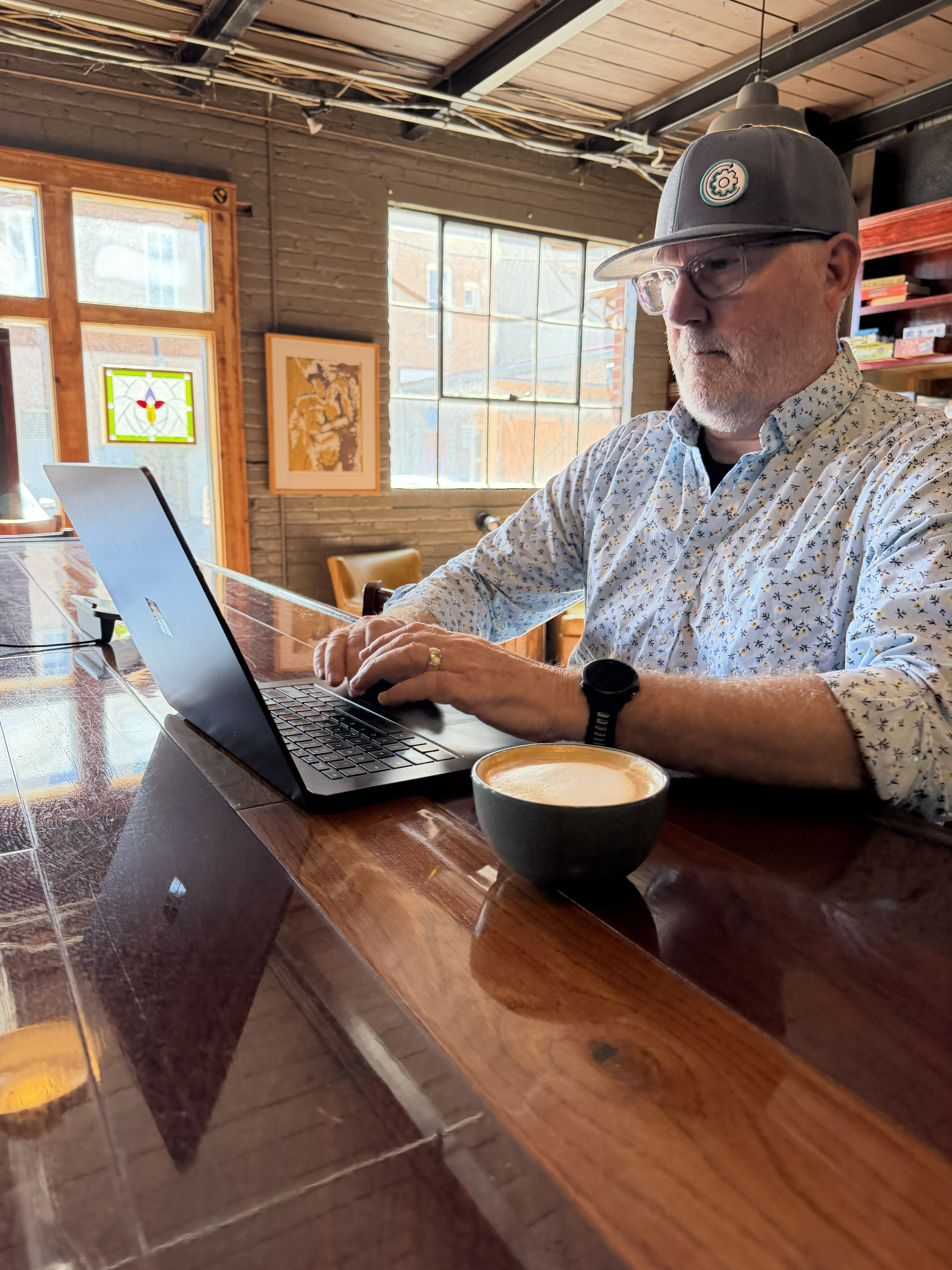 Man with glasses and a gray baseball cap sitting at a wooden table, working on a laptop with a coffee cup in the foreground, inside a cozy cafe with brick walls, large windows, and art on the walls.