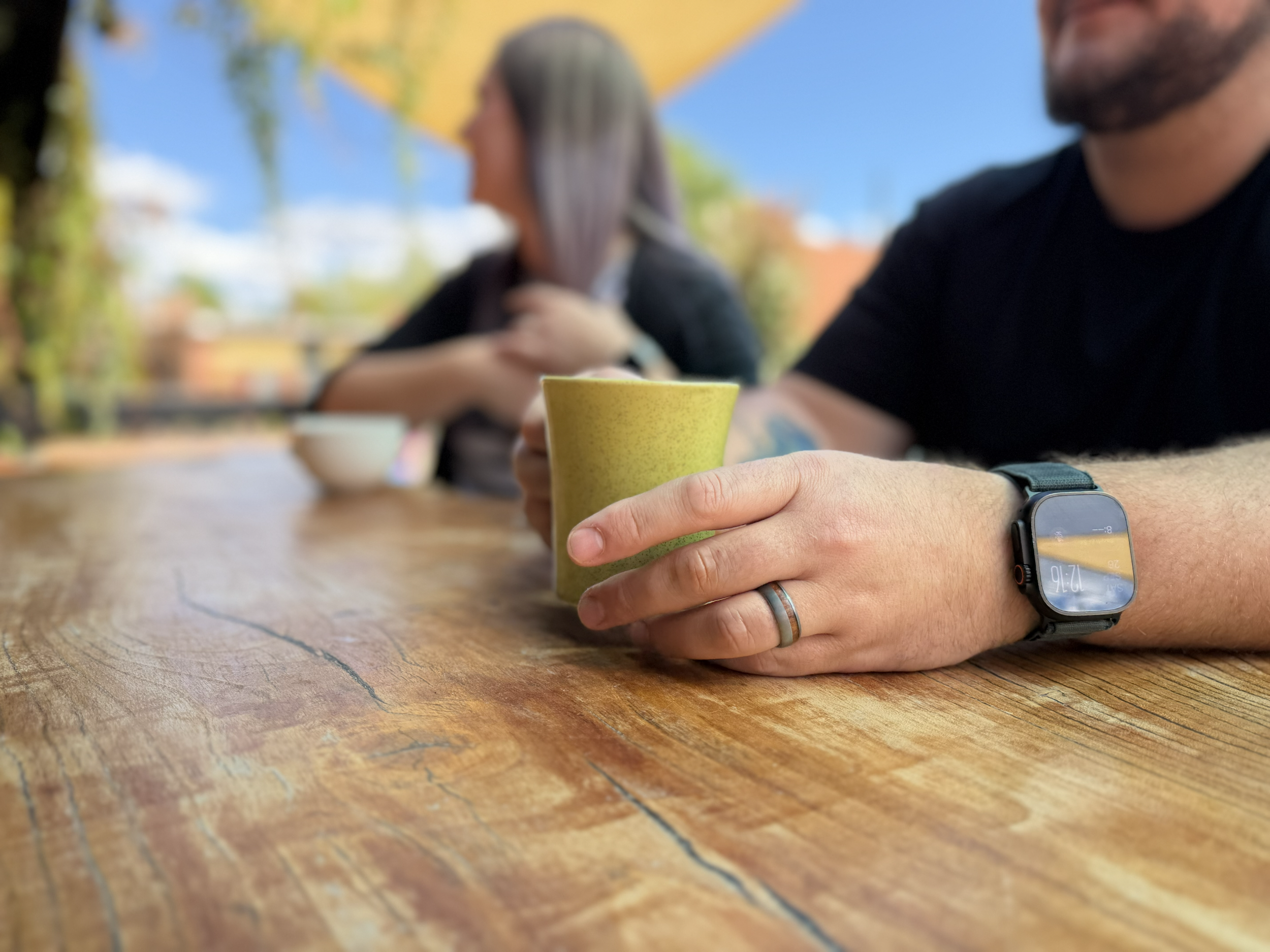 Close-up of a person's hand holding a yellow ceramic mug on a wooden table. The person is wearing a smartwatch and rings. In the blurred background, there are two people sitting outdoors under a yellow umbrella, with trees and a blue sky.