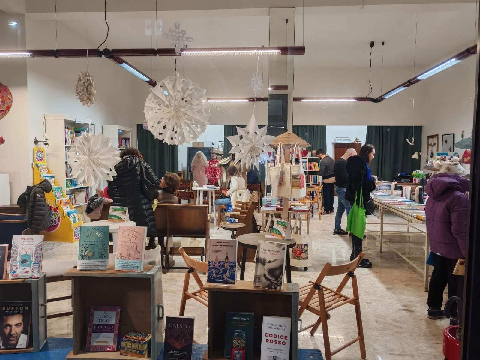 Interno di una libreria o stand di libri con diverse persone che sfogliano e comprano libri. Decorazioni natalizie pendono dal soffitto.