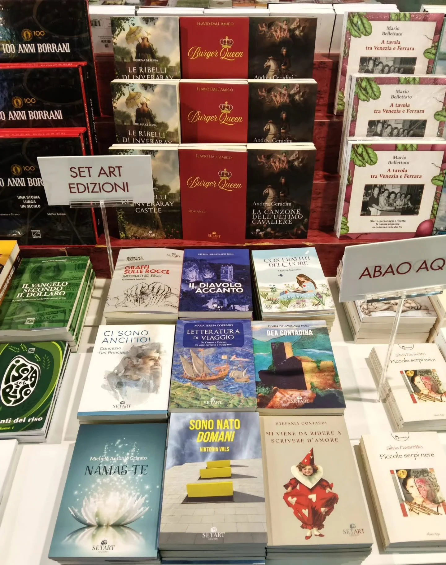 Diversi libri in esposizione in una libreria, con un cartello che dice 'Set Art Edizioni'. Al centro, libri con copertine rosse, nere e blu, tra cui titoli come 'Burger Queen', 'Le Ribelli diInveraray Castle' e altri; sullo sfondo altri scaffali con libri e cartelli.