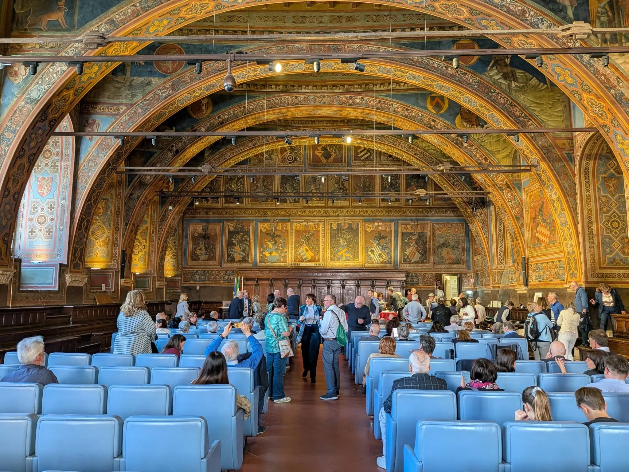 Interno di una grande sala decorata con affreschi sui soffitti e retro del palco, con molte persone sedute e in piedi, alcune scattando foto, in attesa di un evento.