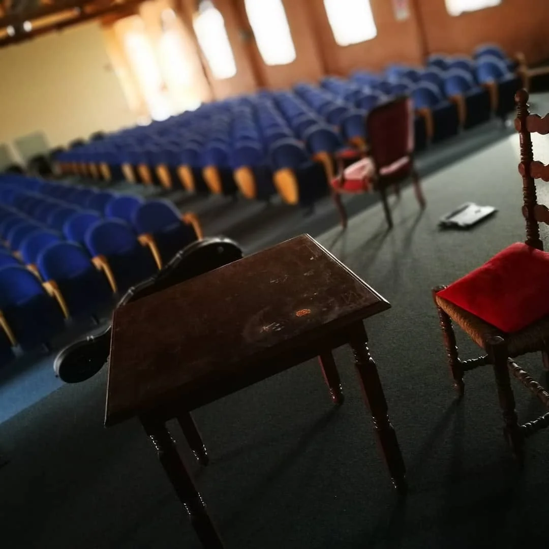Sala vuota con file di sedie blu e due sedie con cuscini rossi e un tavolo in primo piano.