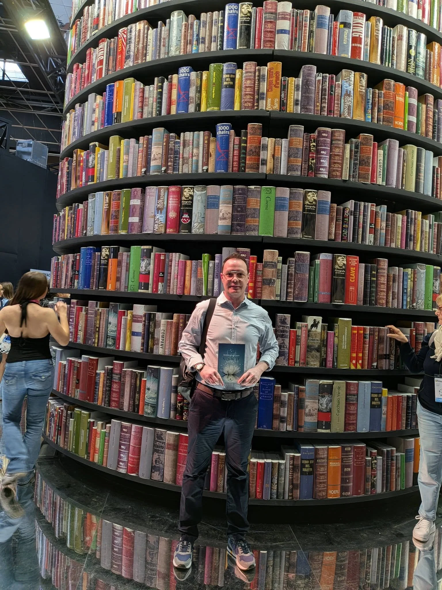 Una persona con camicia azzurra, pantaloni neri e zaino tiene un libro davanti a una grande scaffalatura circolare piena di libri in libreria. Ci sono altre persone intorno, tra cui una donna con camicia senza maniche e una donna con cappotto bianco.