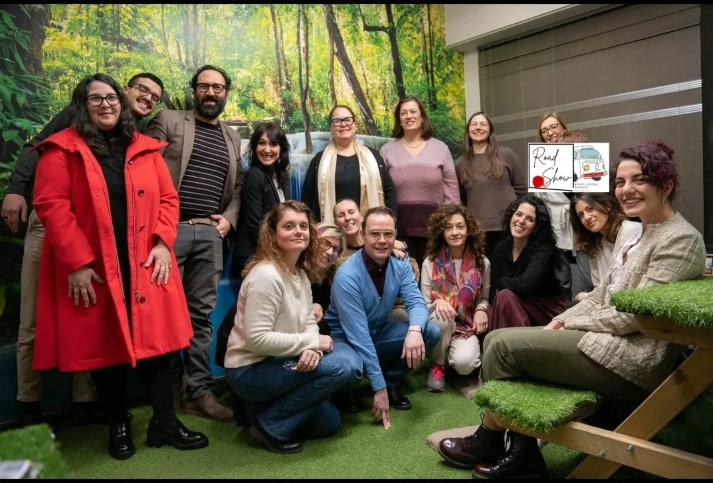 Gruppo di persone che posano per una foto in un ambiente interno con uno sfondo di alberi e natura. L'atmosfera sembra allegra e amichevole.