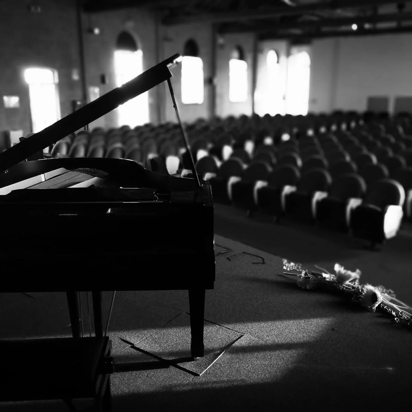 Un pianoforte nero su un palco in un teatro o sala da concerto, con sedie vuote sullo sfondo e decorazioni sul pavimento.