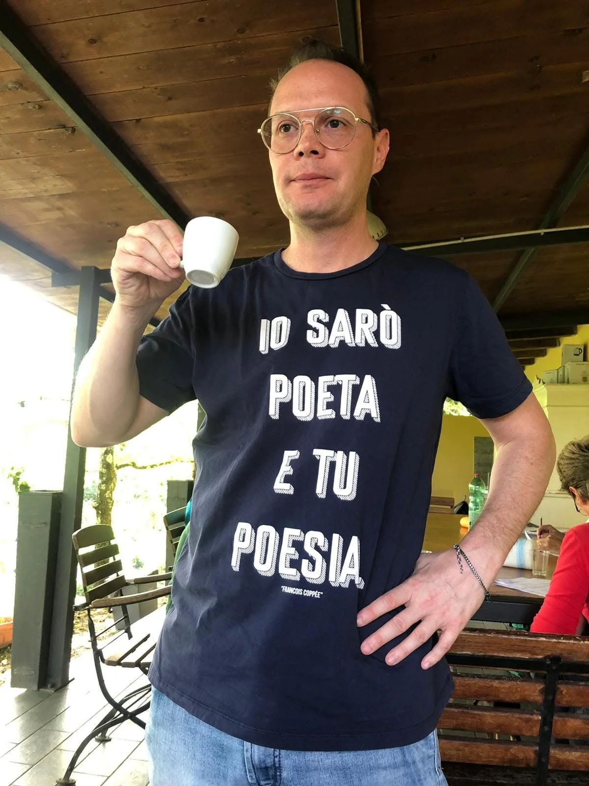 Uomo con occhiali che tiene una tazza bianca, indossa una maglietta con la scritta "Io sarò poeta e tu poesia"