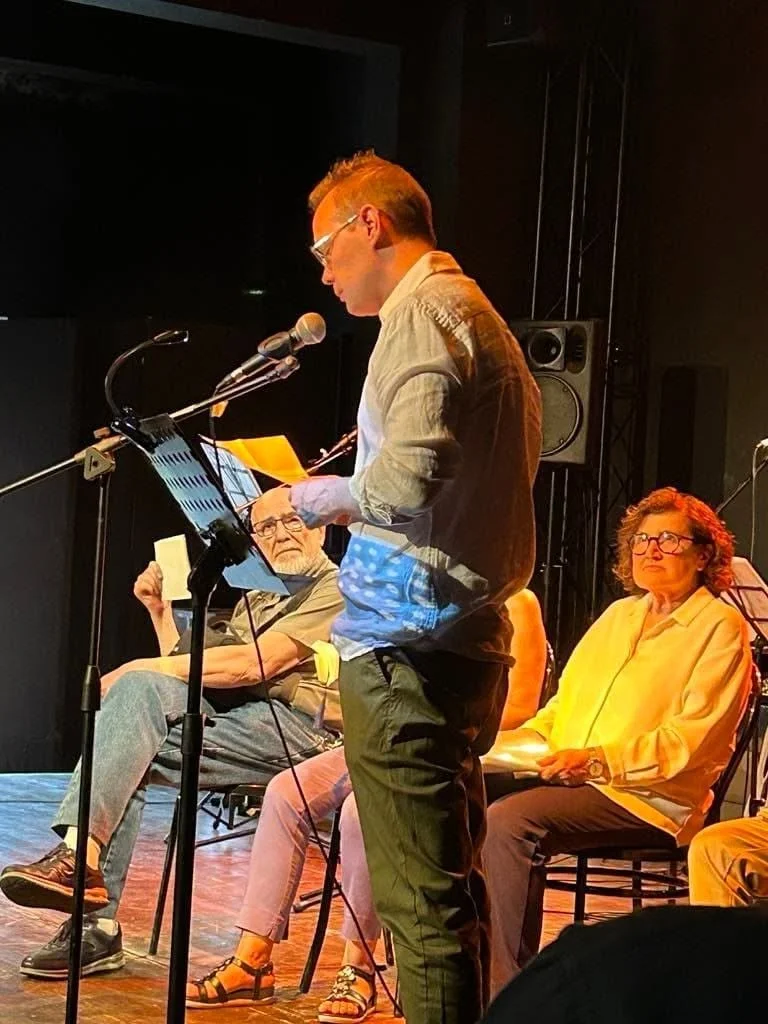 Persona che legge un discorso o una poesia durante un evento sul palco, con altre persone sedute dietro di lui