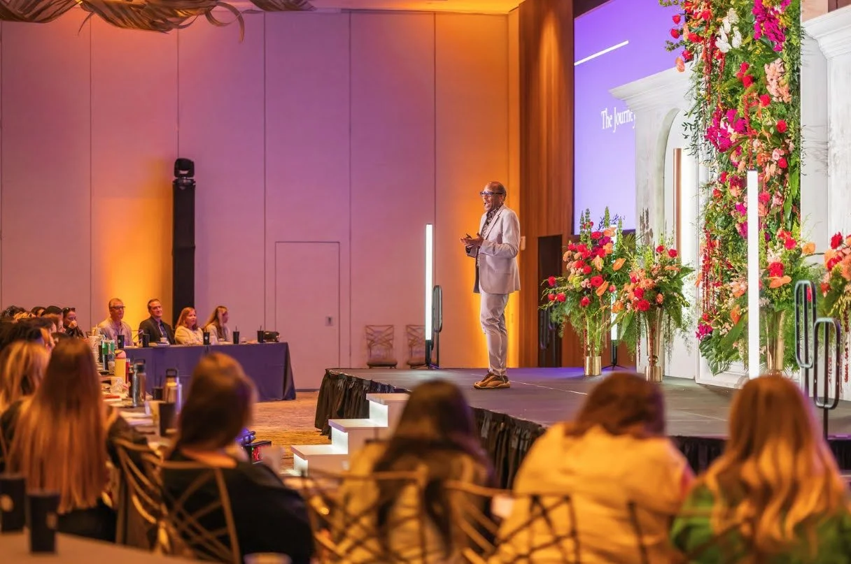 Speaker on stage with floral columns and ambient lighting at a conference in Sarasota