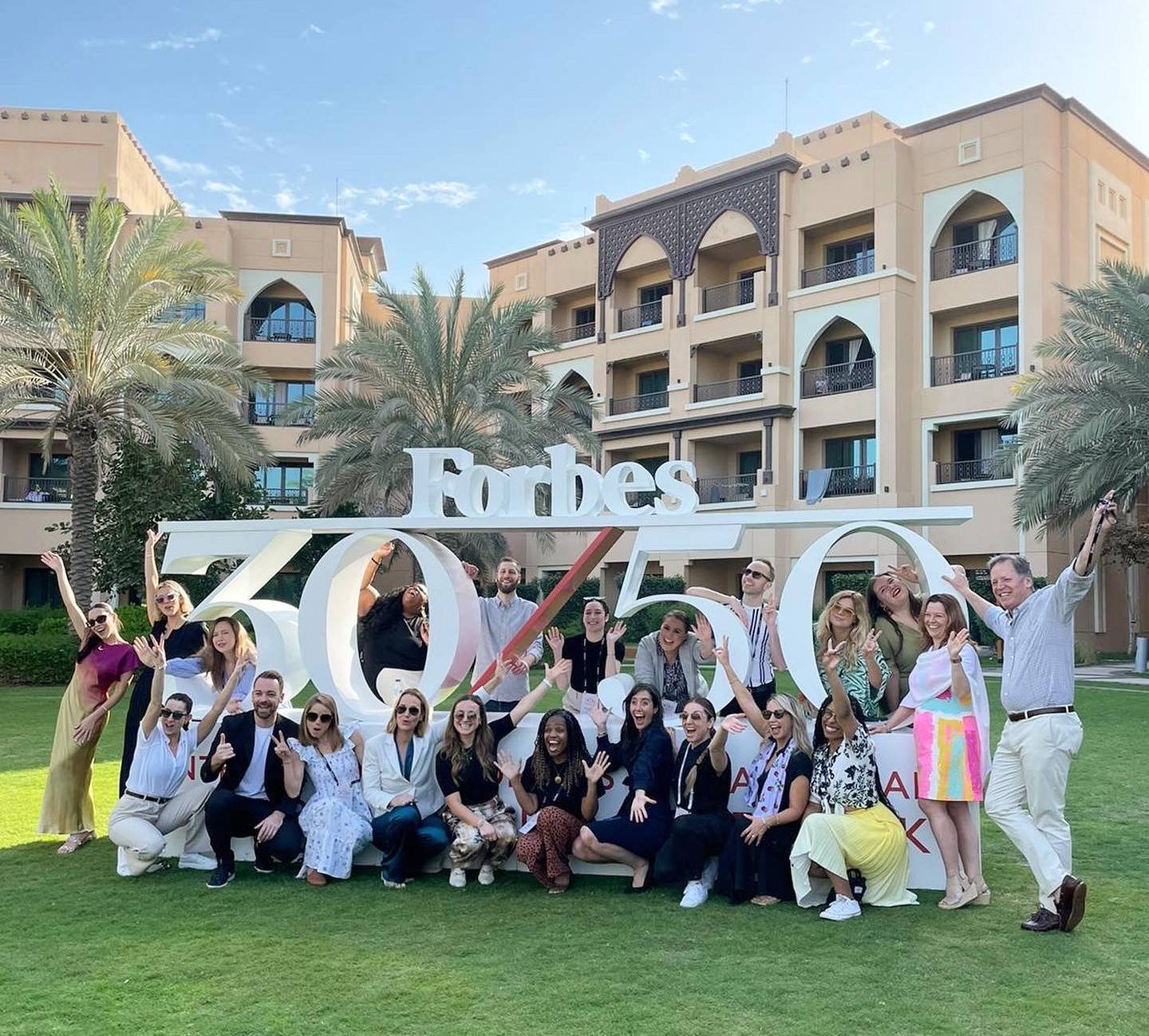 A group of people posing in front of a large 'Forbes 30 Under 30' sign outdoors on a grassy area with palm trees and a multi-story beige building in the background. The group is smiling, waving, and making celebratory gestures on a sunny day.