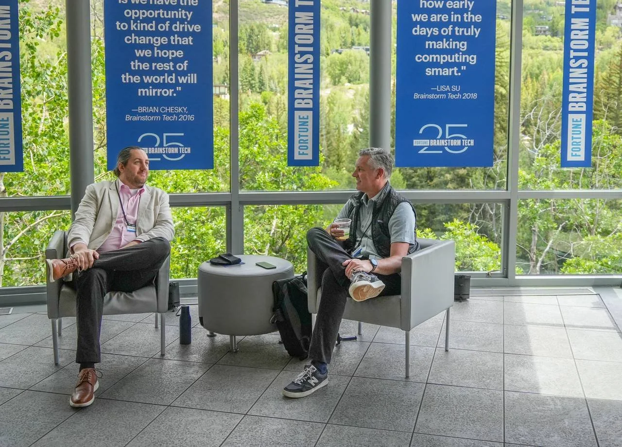 Fortune Brainstorm Tech 2025 attendees talk while seated in lounge chairs near windows hung with event banners.