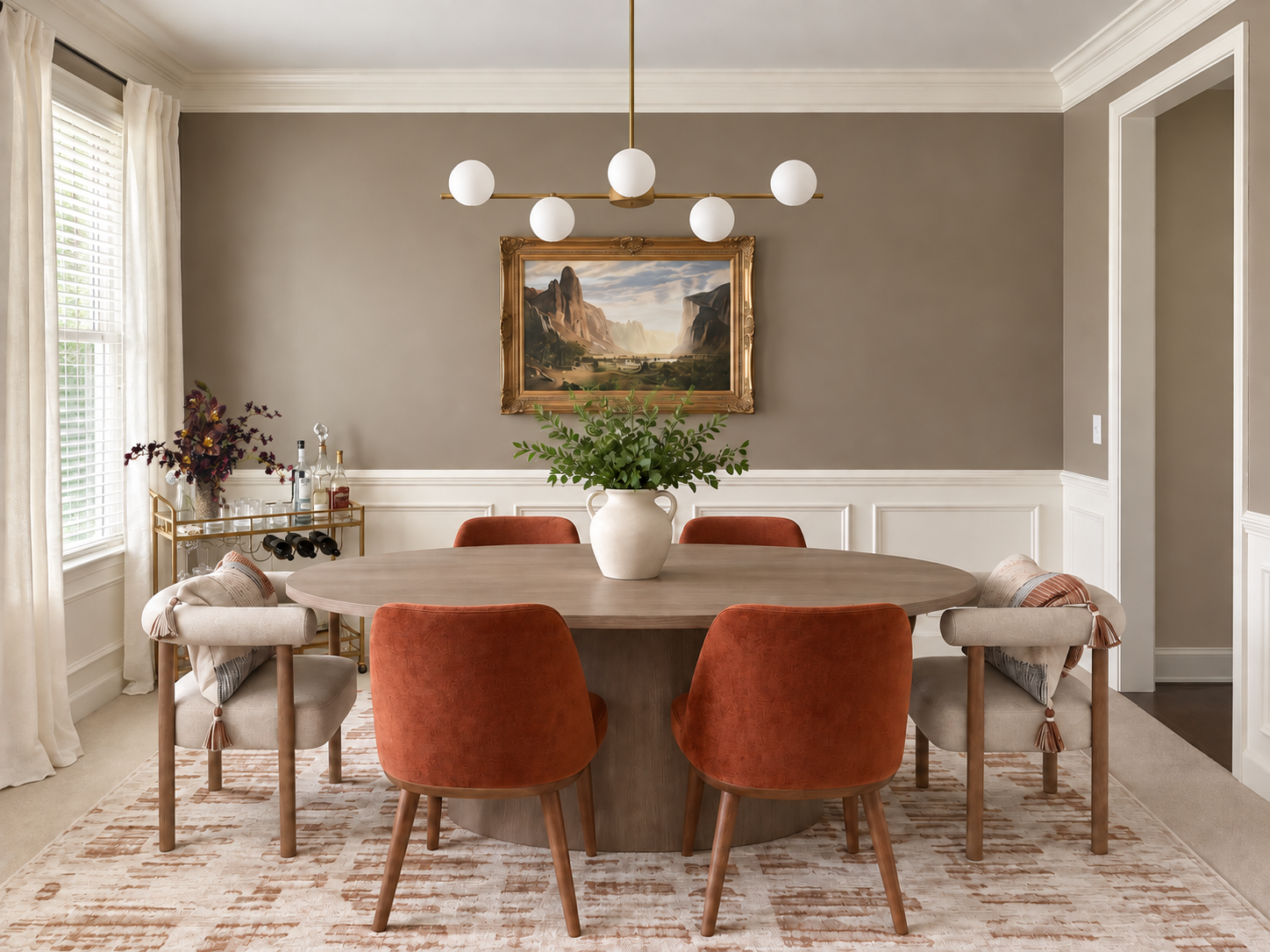 A dining room with a round wooden table surrounded by six chairs, some with orange upholstery and some with beige and tassel accents. A large vase with green foliage sits in the center of the table. A painting of a landscape with mountains hangs on the beige wall above the table. A modern chandelier with six white globes hangs from the ceiling. To the left, a window with white curtains and a bar cart with bottles and glasses are visible.
