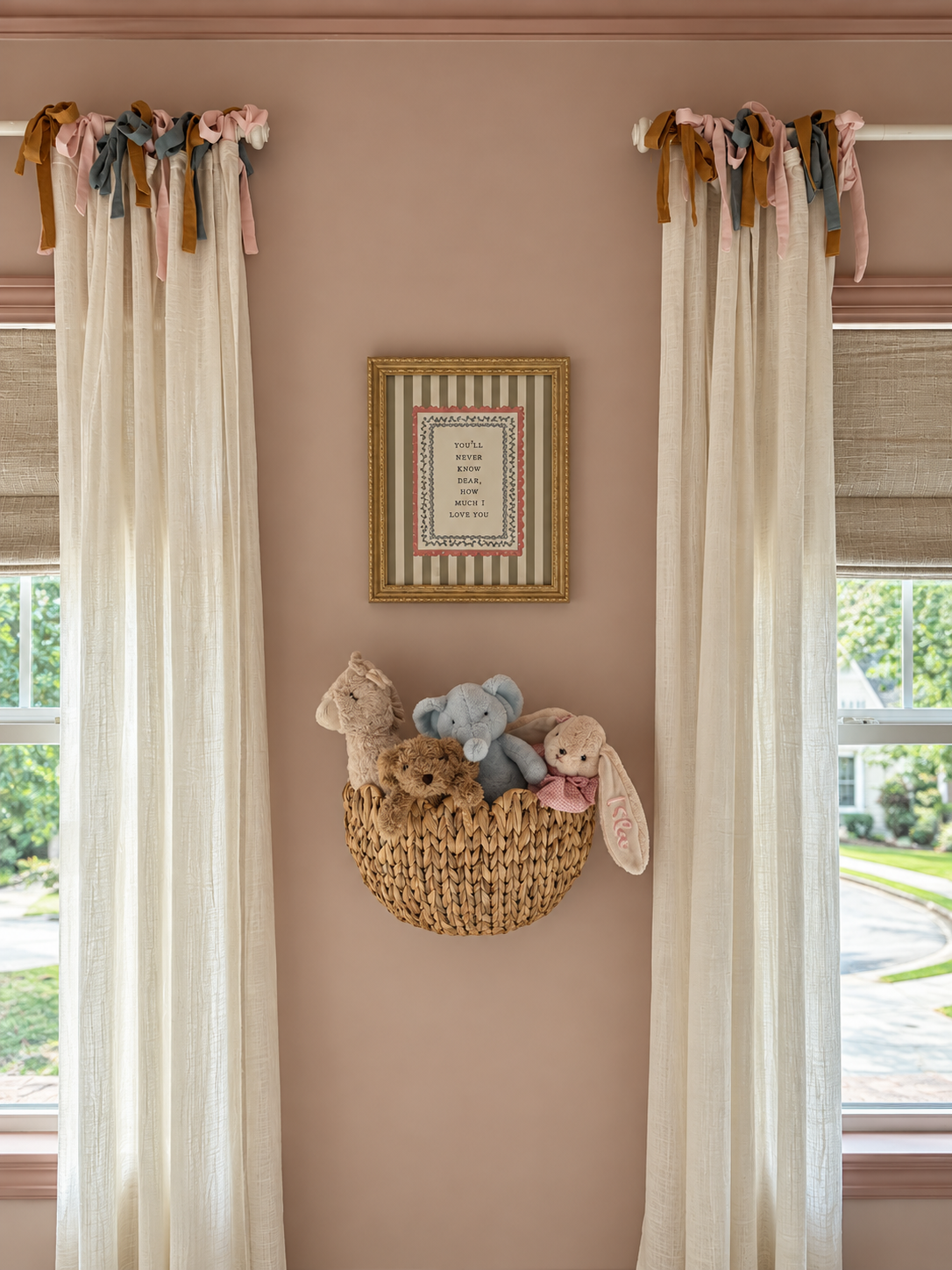 A wall with two cream-colored curtains decorated with colorful bows at the top. Between the curtains is a framed quote on the wall. Below the quote, a wicker wall basket holds plush stuffed animals, including a beige bear, a gray elephant, a small brown bear, and a white bunny with pink ears.