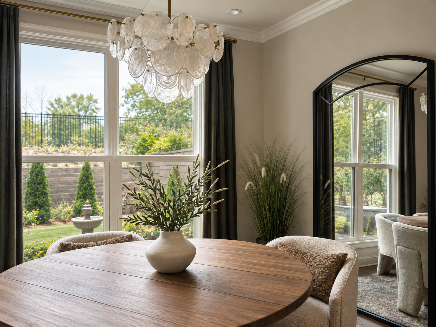 Dining area with a wooden table, a white vase with greenery, large windows with dark curtains, and a decorative chandelier hanging from the ceiling.