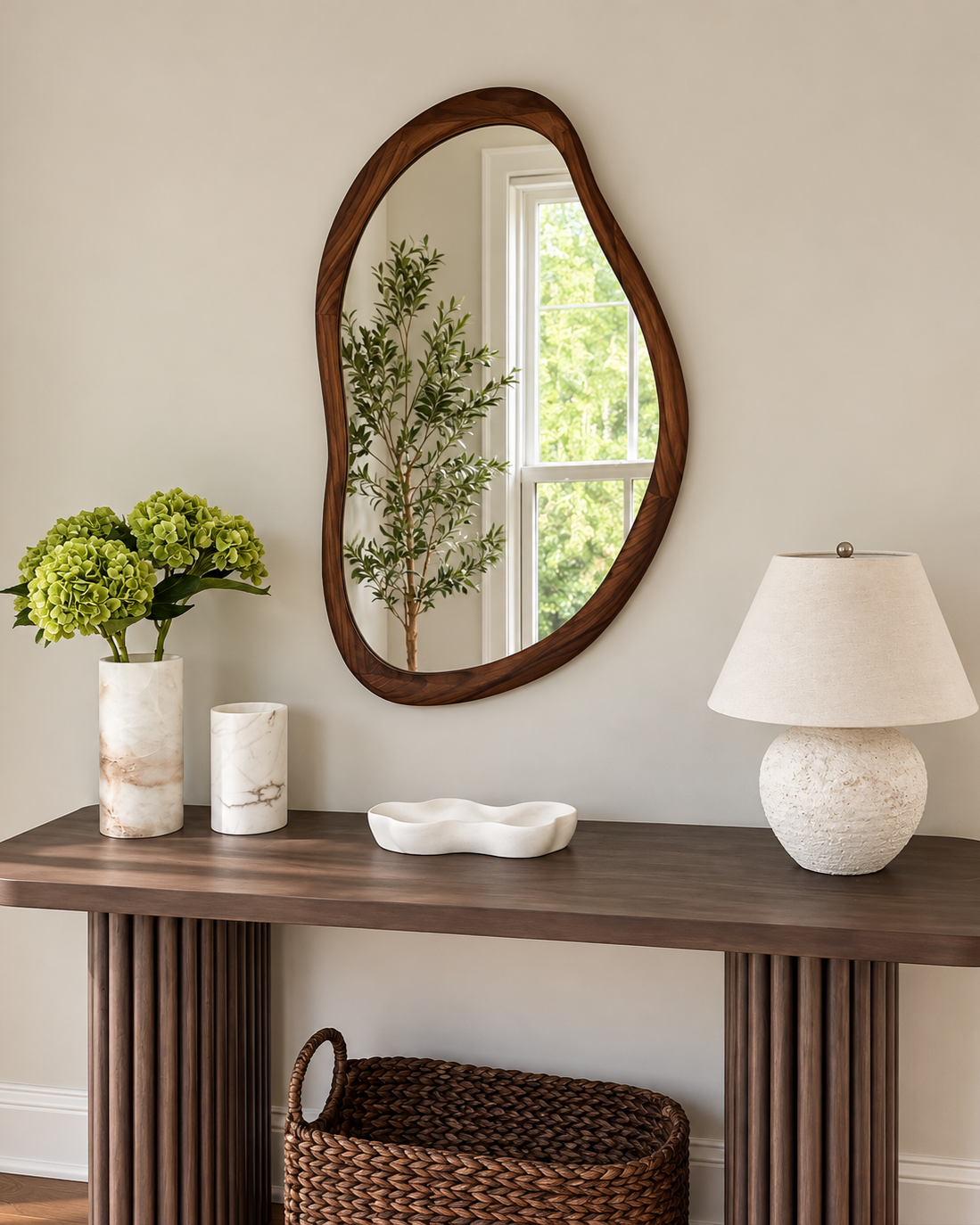 Decorative wooden framed mirror on a wall, above a wooden console table with green hydrangeas in a marble vase, a white ceramic dish, and a textured white table lamp with a umbrella shape shade. A woven basket is beneath the table.