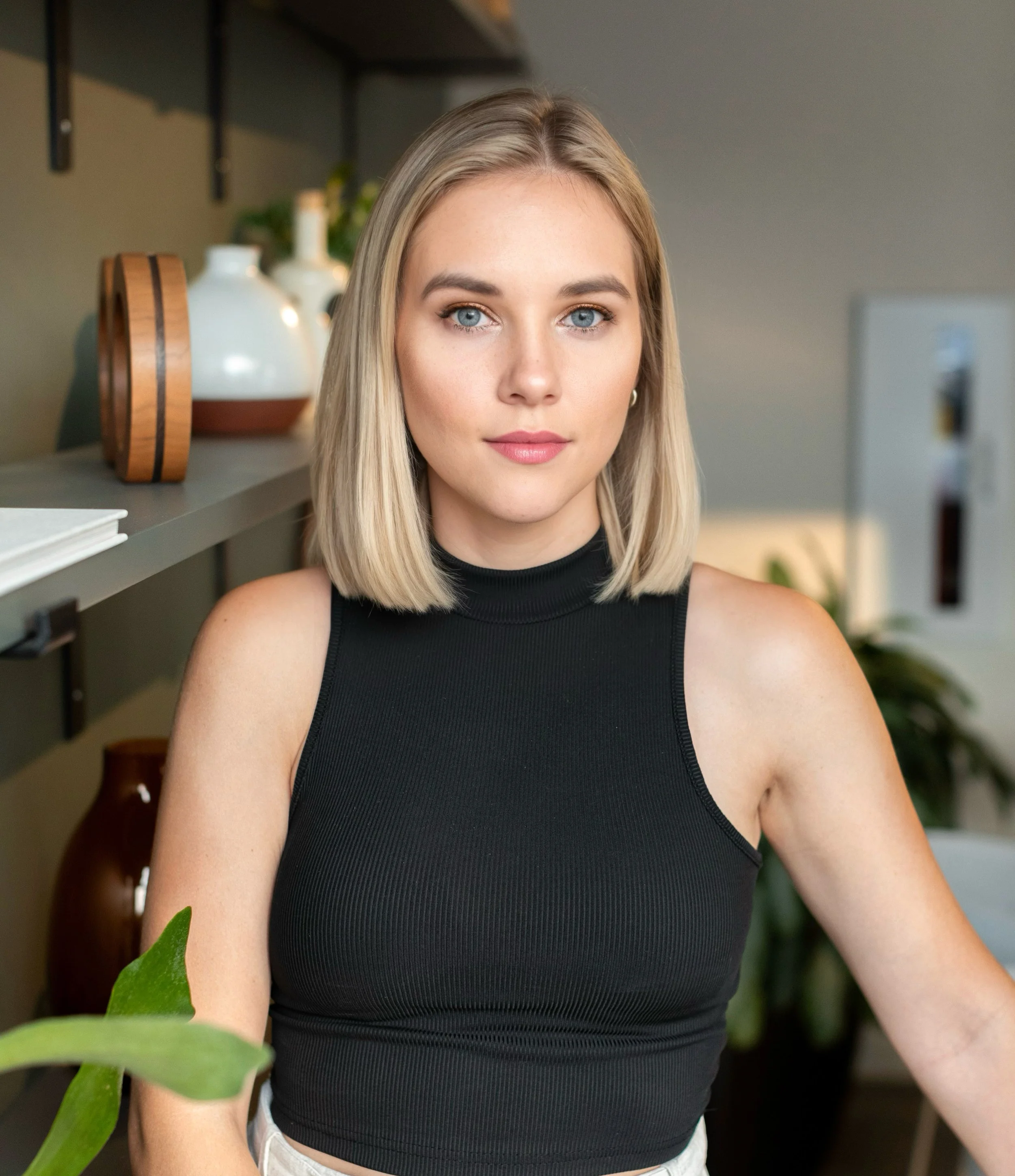 Close-up of a young woman with blonde hair, blue eyes, and light skin, wearing a black sleeveless top, standing indoors near a shelf with decorative vases, with a green plant in the foreground.