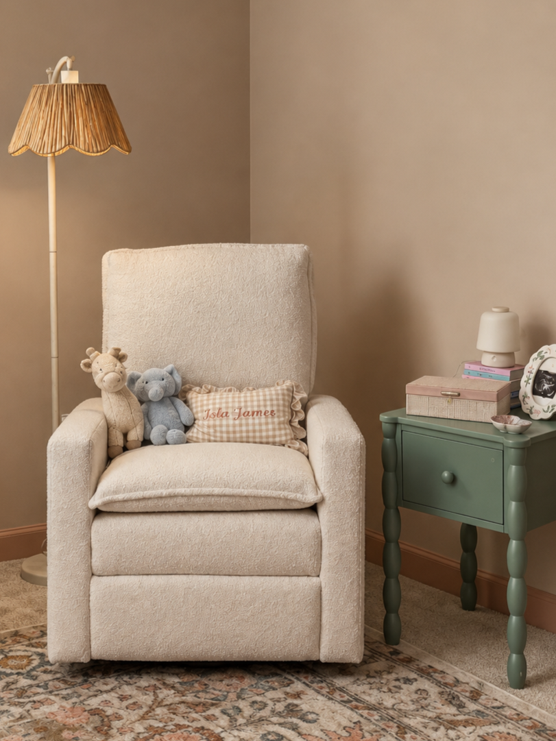 A cozy corner with a cream armchair, plush teddy bears on the cushion, a pink ruffled pillow with embroidered text, a green side table with books, a white lamp, and a floor lamp with a woven lampshade, on a patterned rug.
