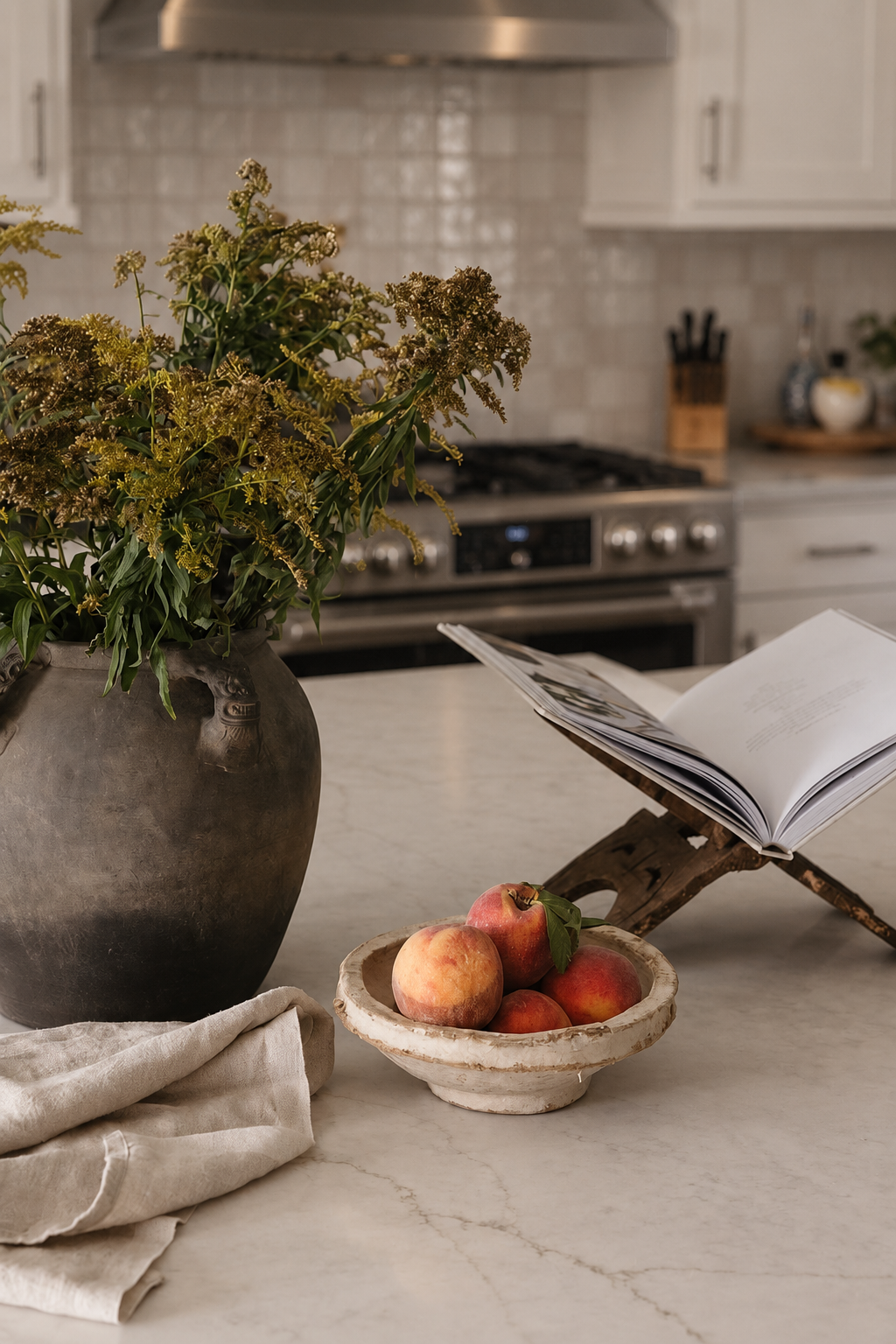 A kitchen countertop with a large vase of greenery, a bowl of peaches, and an open book on a wooden stand, with kitchen appliances and utensils in the background.