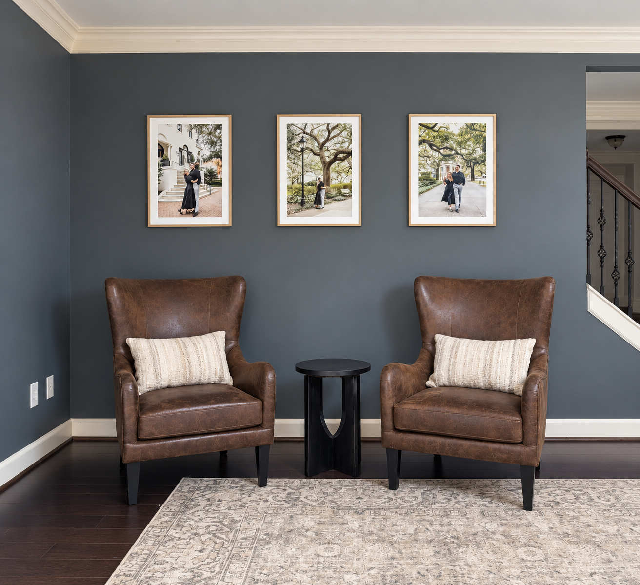 Living room with dark gray walls, two brown leather armchairs with beige pillows, a small black side table between them, a patterned rug, three framed photographs of a couple outdoors on the wall, and a staircase with a railing visible in the background.