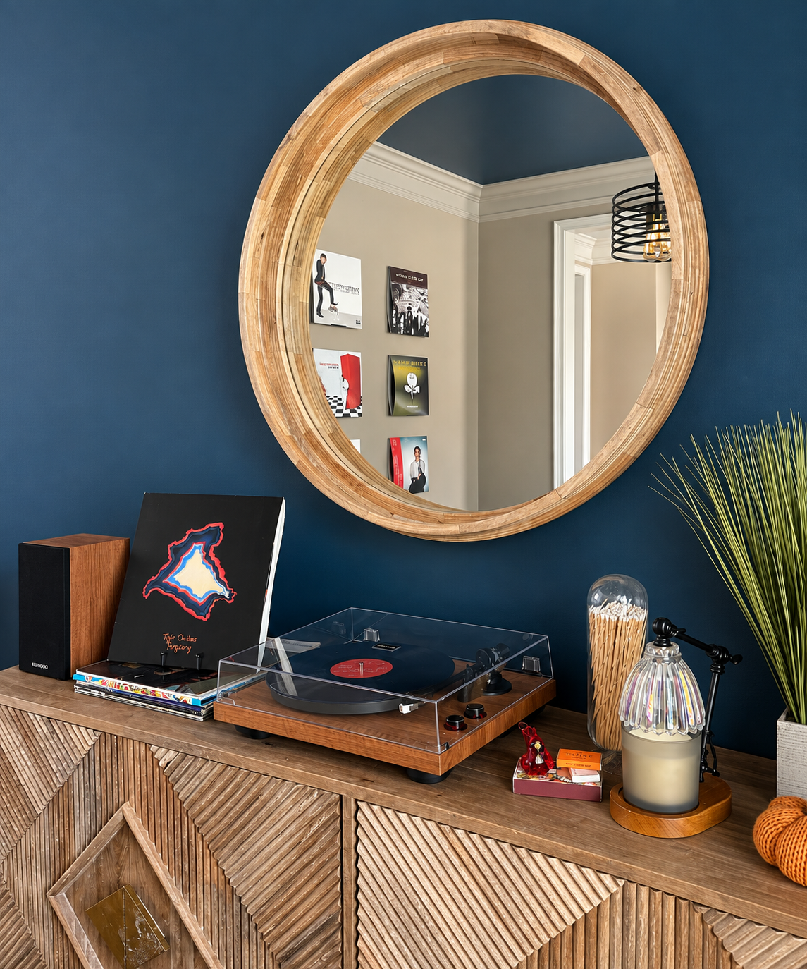 Interior of a room with a wooden sideboard, decorated with a record player, vinyl records, a speaker, a lamp with a clear shade, a potted plant, and various decorative items. A large round mirror with a wooden frame is mounted on a blue wall, reflecting part of the room with posters on a beige wall and a doorway.