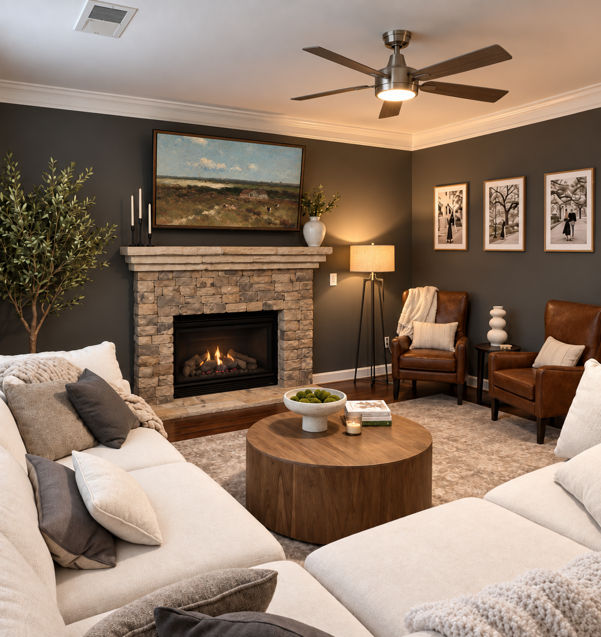 Living room with a stone fireplace, a ceiling fan, a large white sofa with pillows, a wooden coffee table with a bowl of moss and books, brown leather chairs, a tall floor lamp, framed artwork on dark walls, and decorative objects.