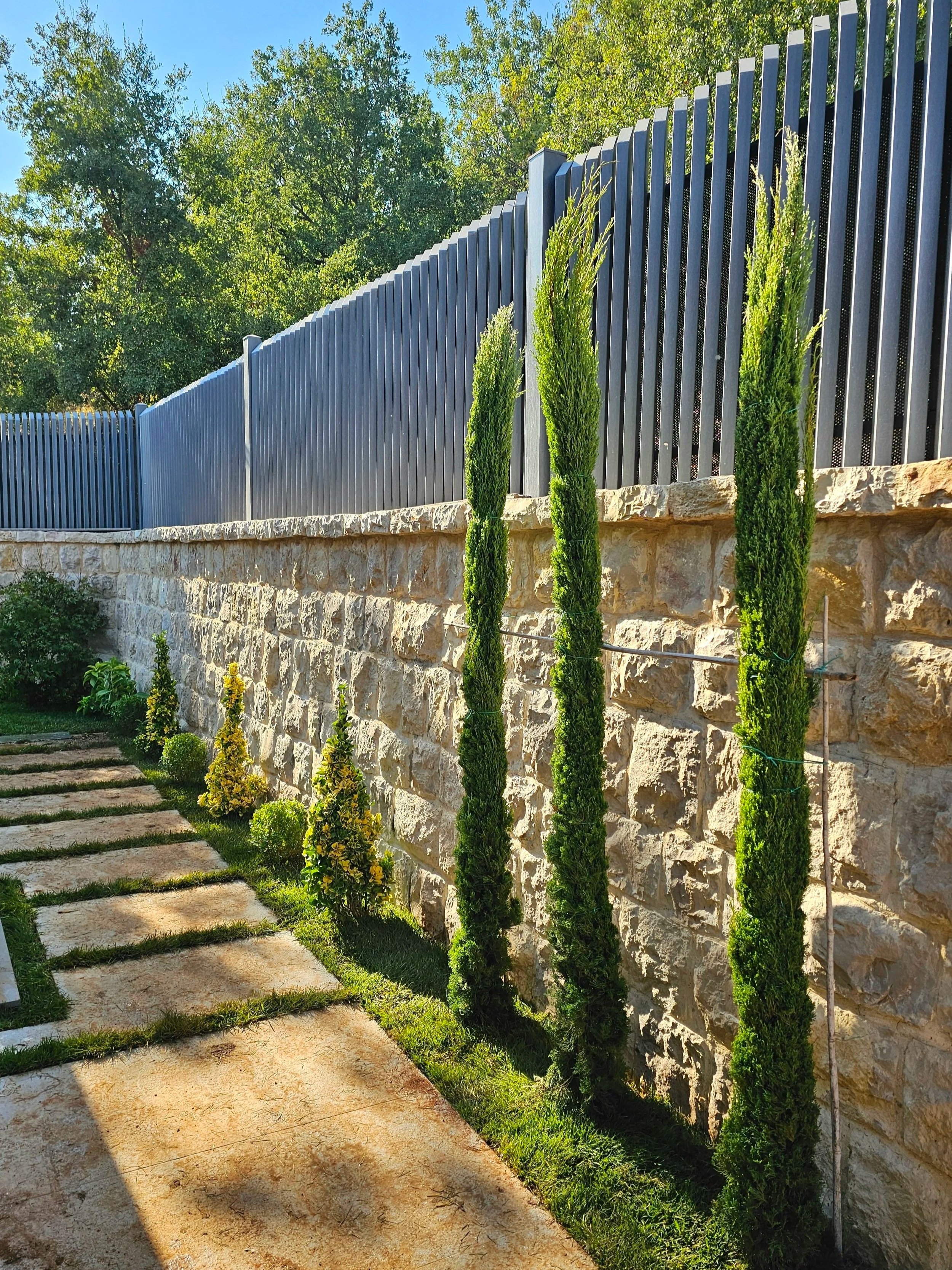 A landscaped outdoor area with a stone retaining wall, a modern dark gray metal fence, and tall green trees and plants against a blue sky.