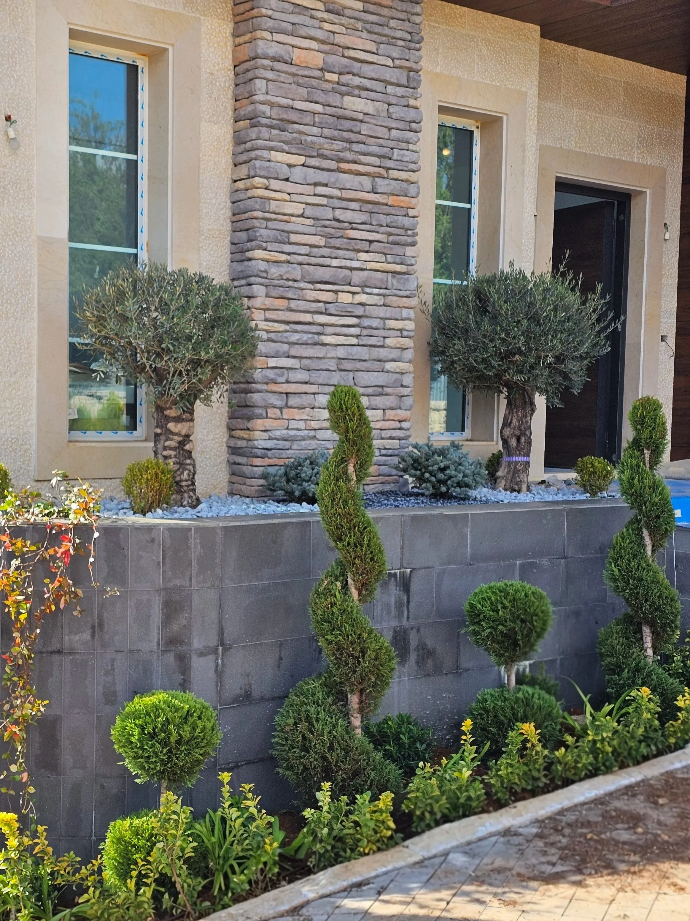 Front view of a modern house with beige and gray exterior walls, stone pillar, and stylish landscaping including shaped shrubs and a low stone wall.