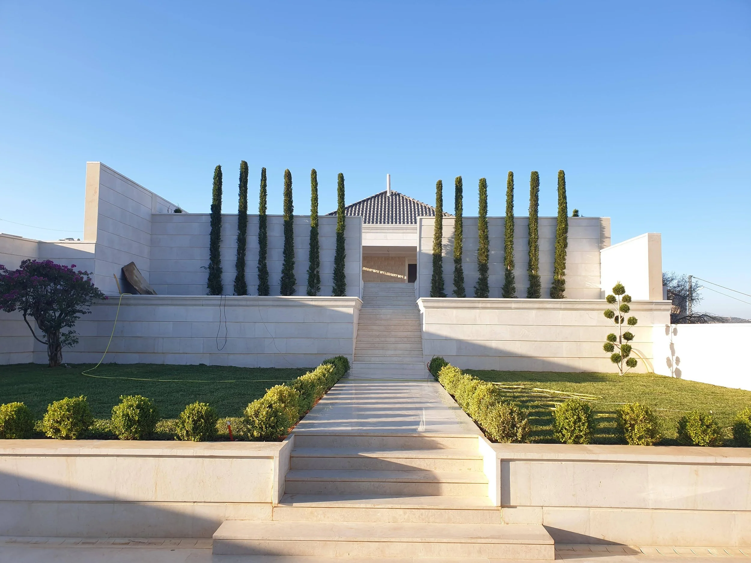 A modern white building with tall, slender trees lining its upper level and a white staircase leading up to the entrance, surrounded by a well-manicured lawn with small bushes and trees, under a clear blue sky.