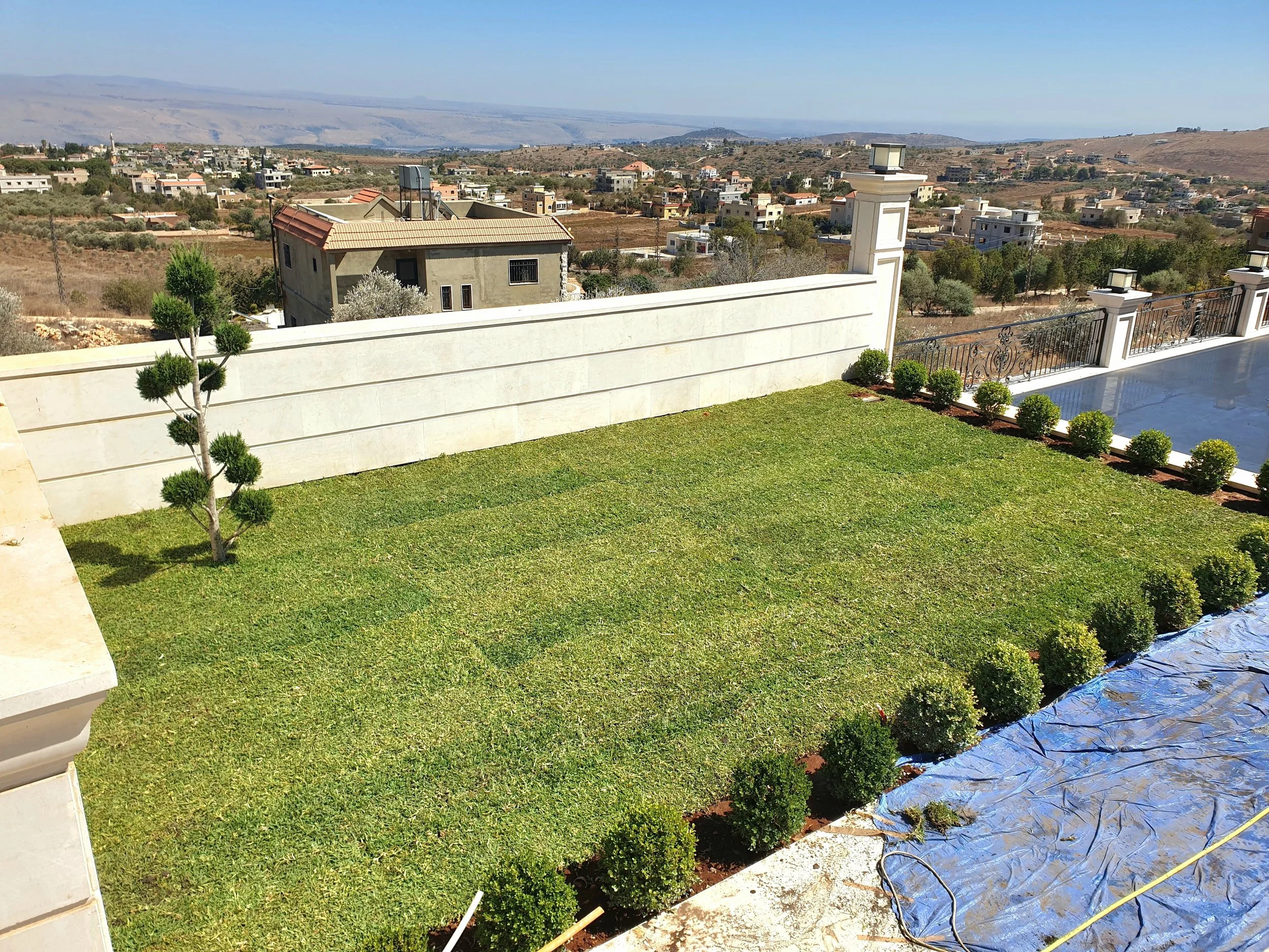 A backyard with a green lawn, small trees, and a white wall with a black wrought iron fence. The background includes a hilly landscape with houses and dry land under a blue sky.