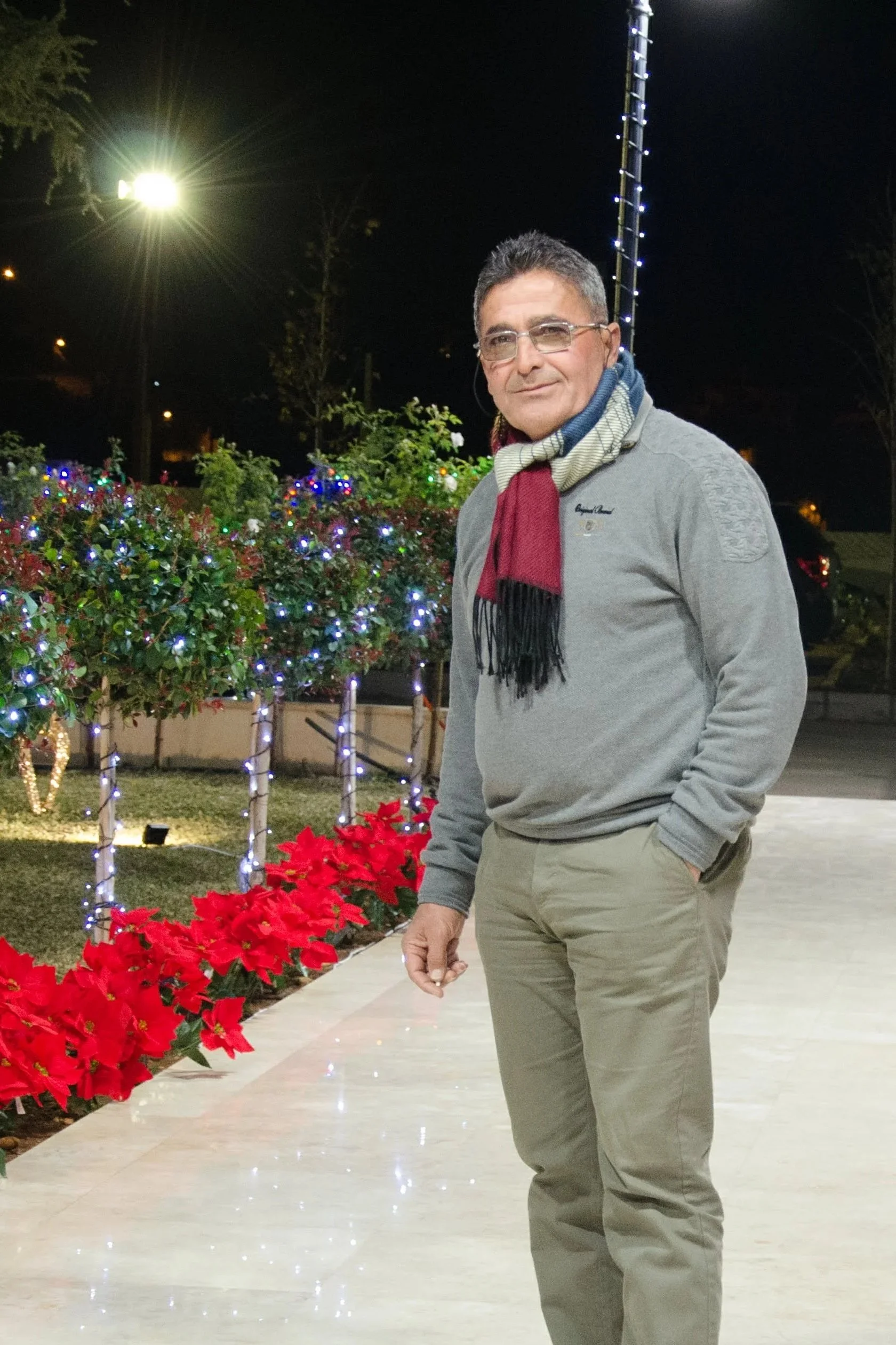 A man with glasses and short hair wearing a gray sweater, colorful scarf, and khaki pants standing outdoors at night with Christmas lights and poinsettias in the background.