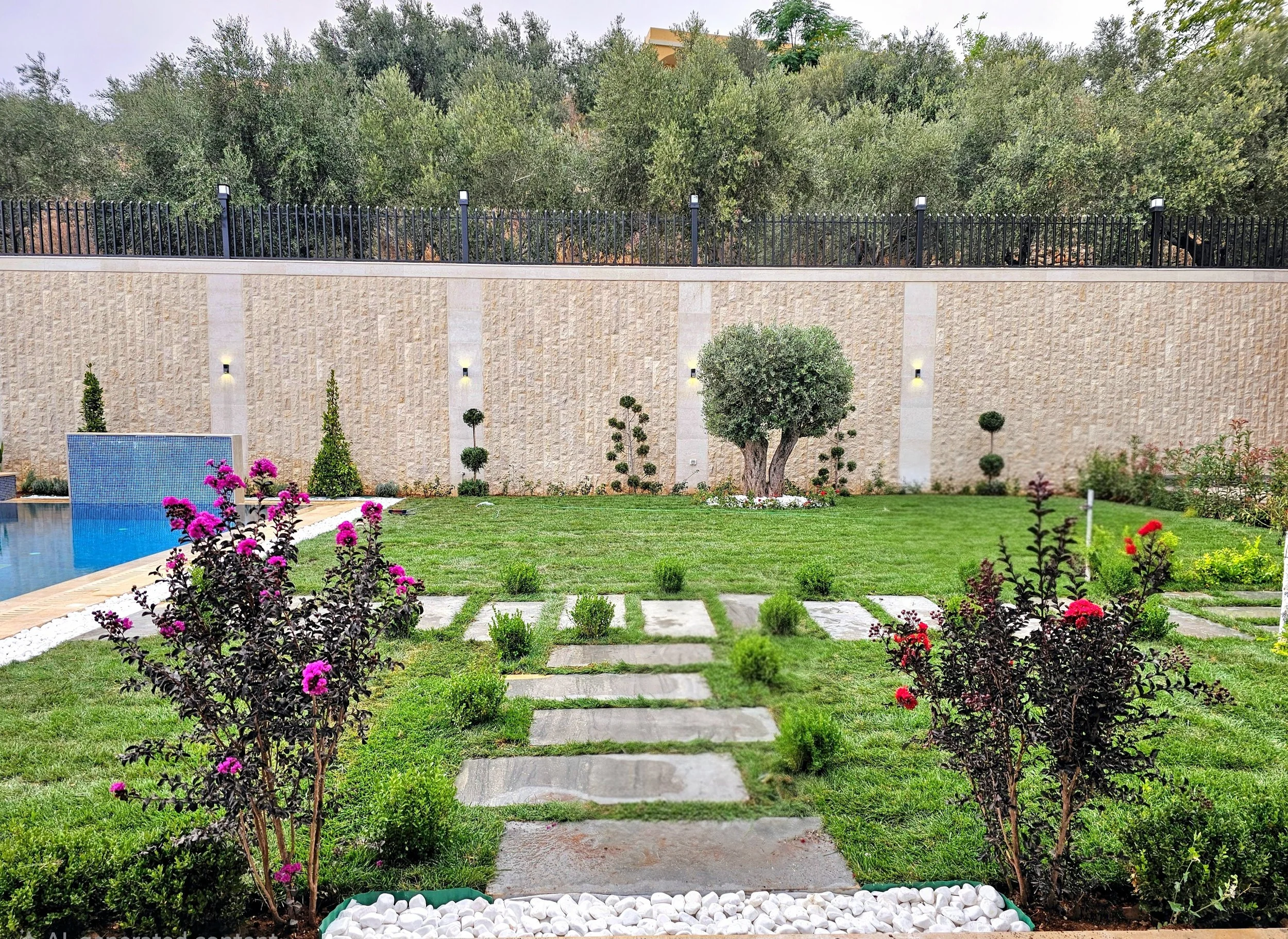 A backyard garden with a stepping stone pathway, colorful flowers, trimmed bushes, a manicured lawn, a beige wall with lights, and trees in the background.