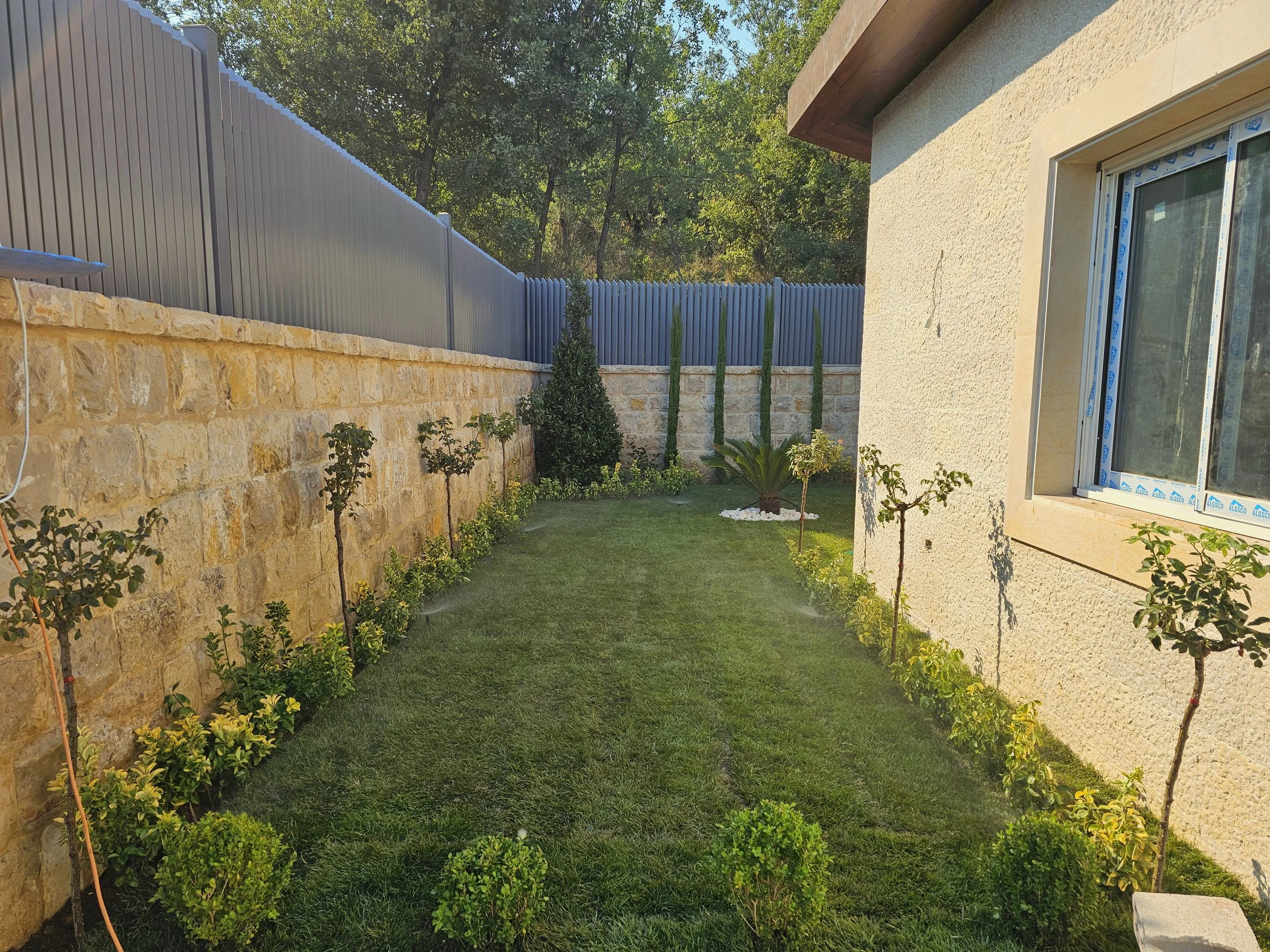 A backyard garden with a stone wall and a beige house wall with a window, a gray fence, small trees, bushes, and a patch of grass