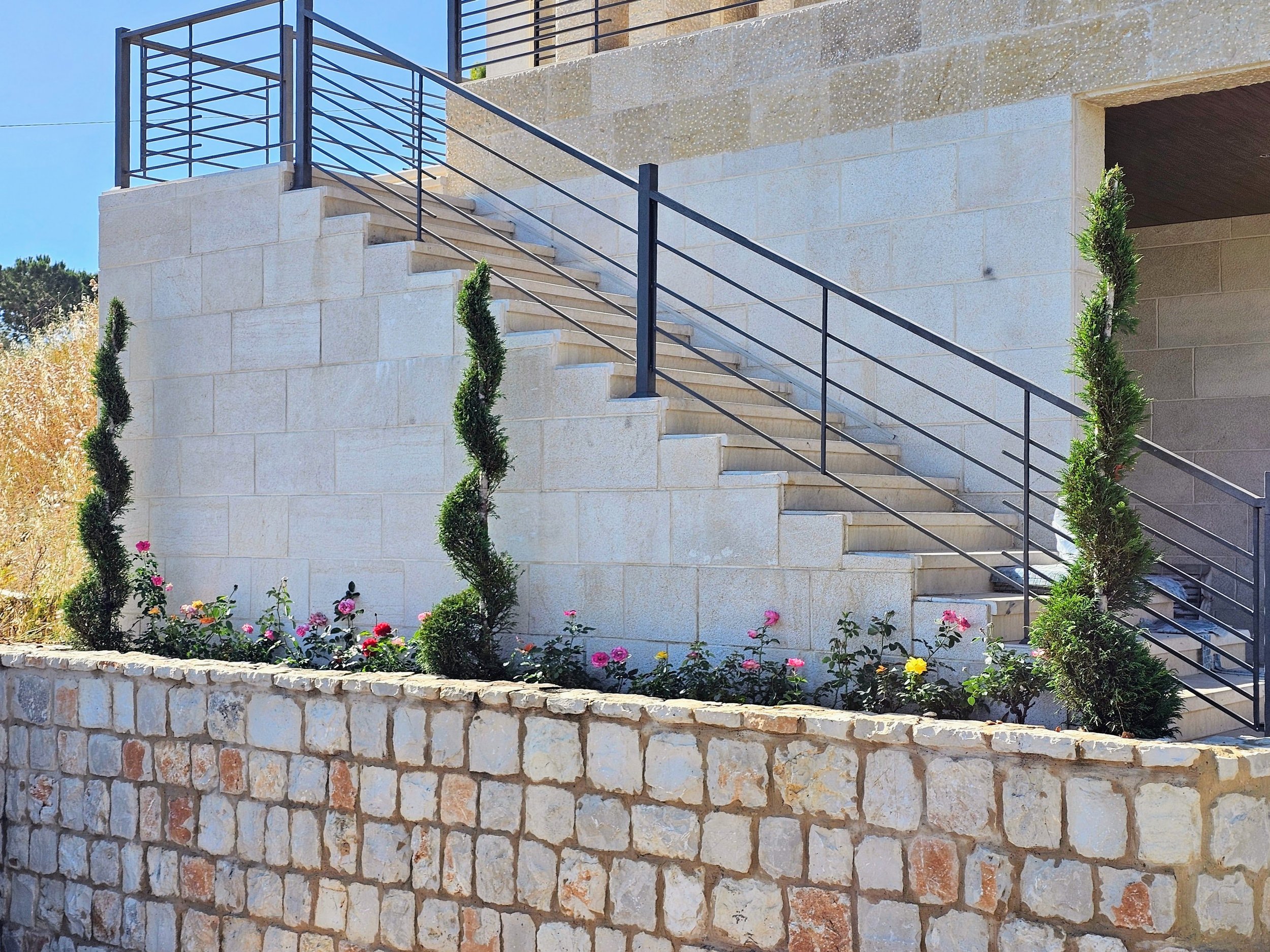 Exterior view of a modern building featuring a stone staircase with black metal railings. The surrounding area has a stone wall planter with green spiral-shaped shrubbery and colorful flowers. The building's upper level has a balcony with a black metal railing. The sky is clear and blue.