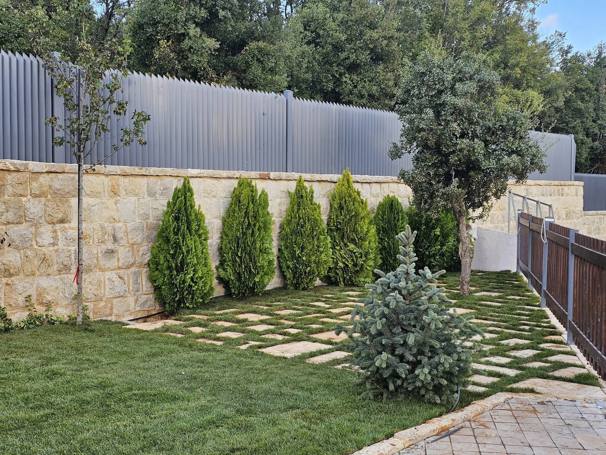 A backyard with a stone wall, green bushes, a small tree, and a tall metal fence with trees and blue sky in the background.