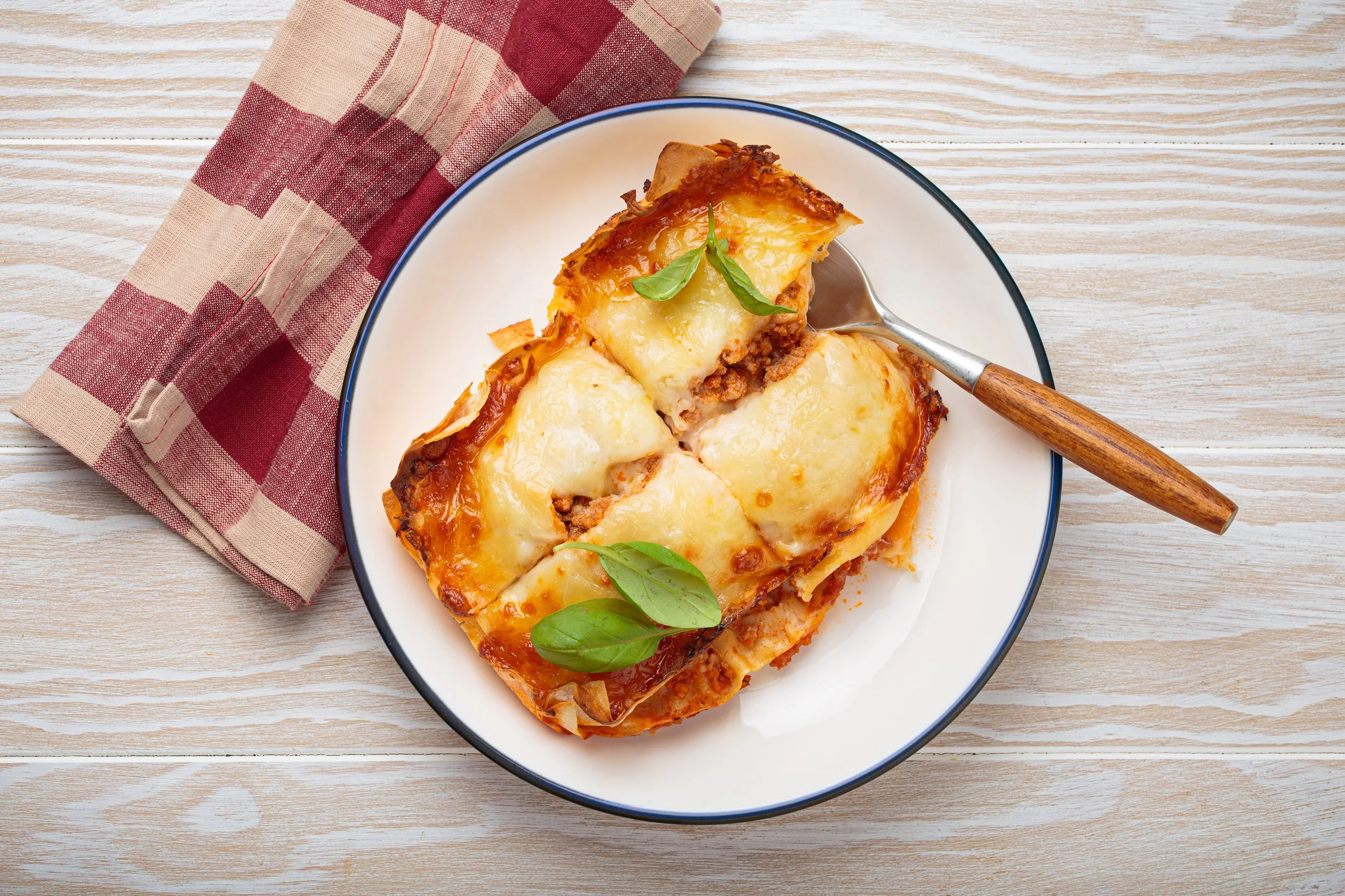 A plate of baked lasagna topped with melted cheese and fresh basil leaves, placed on a wooden surface with a red checkered cloth nearby.