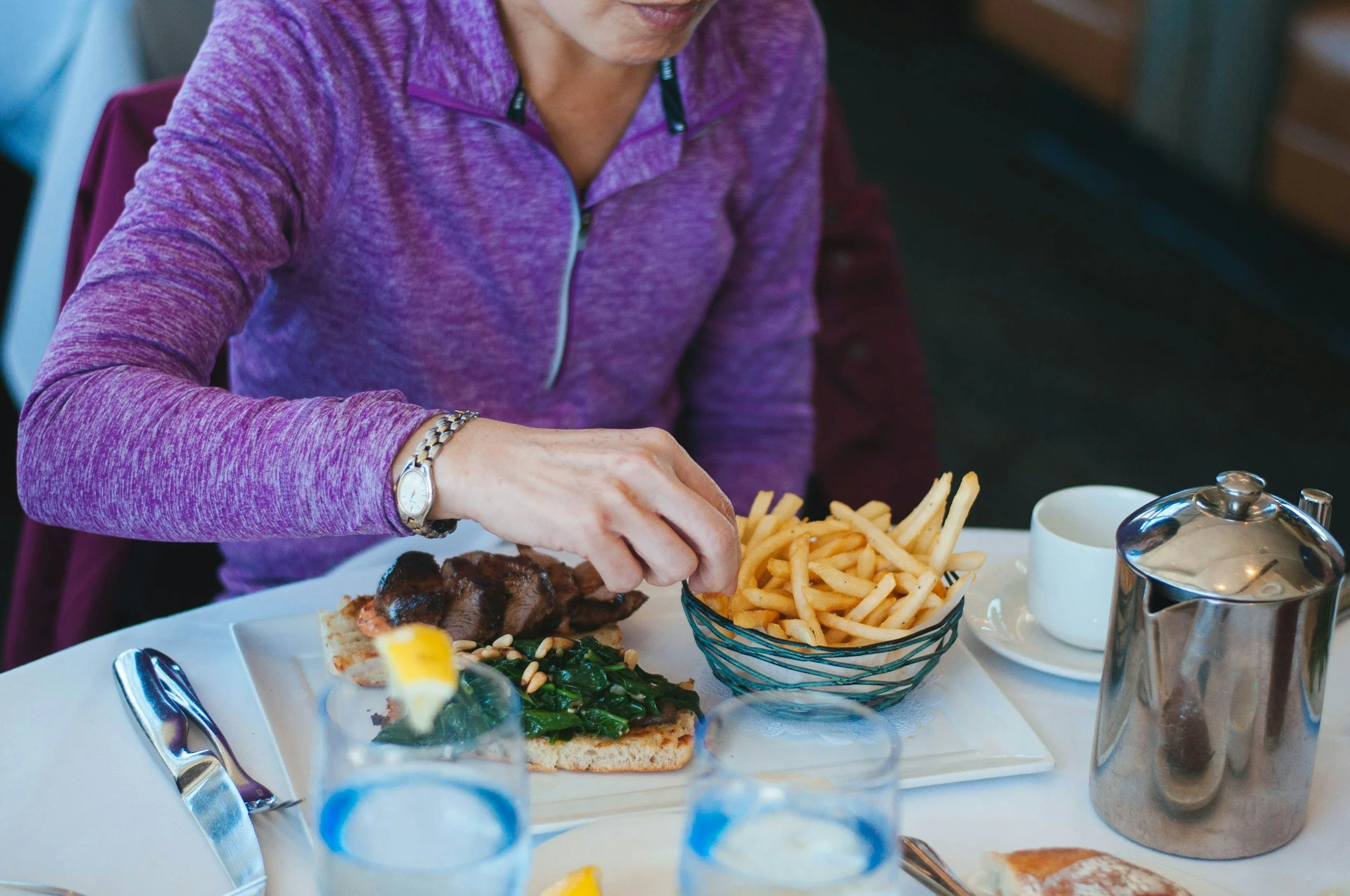 A woman dressed in a purple zip-up jacket is seated at a table with a plate of food. The plate contains French fries, a piece of bread with greens and meat, and a small bowl of sauce. In front of her, there are two blue-topped glasses and a silver teapot. The woman is wearing a wristwatch and gold bracelet.