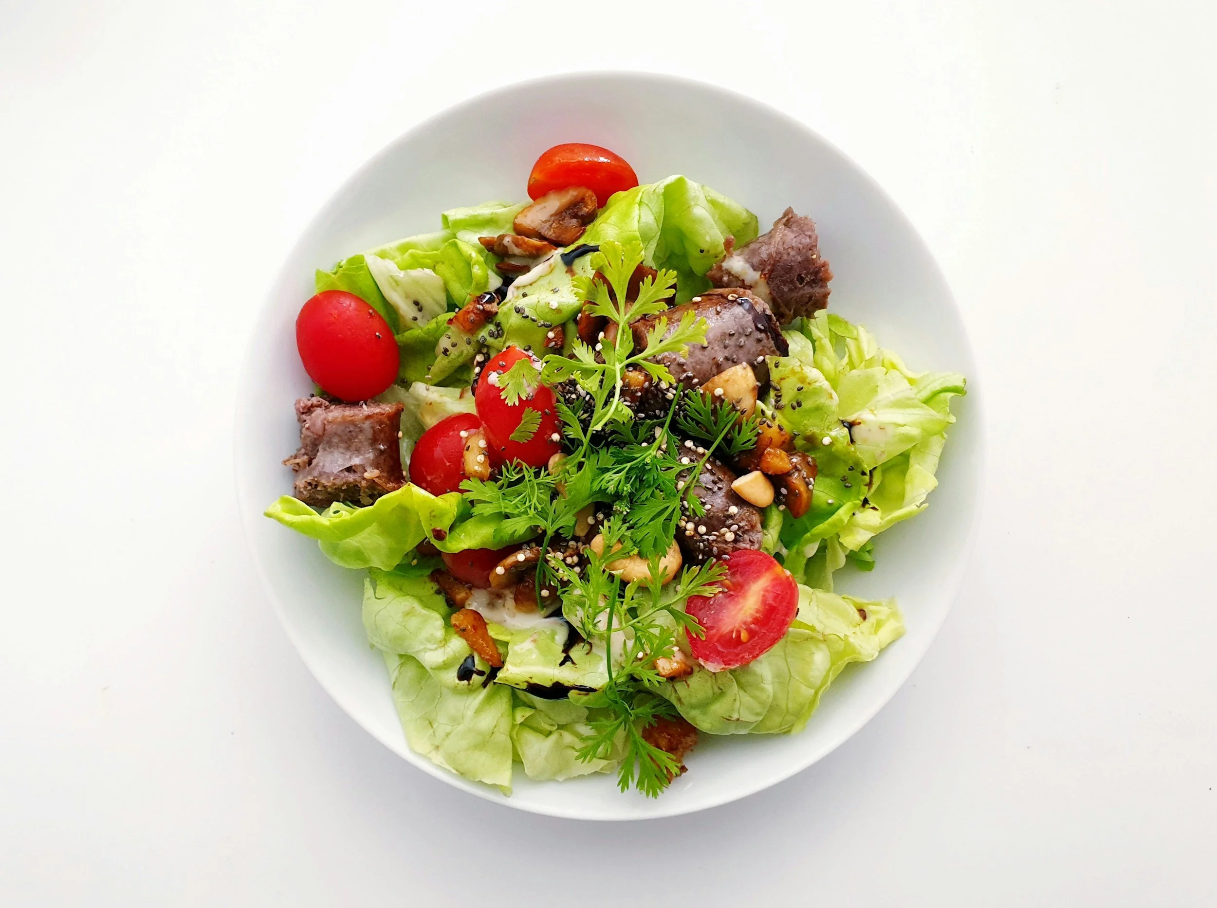 A white plate with a fresh salad consisting of lettuce, cherry tomatoes, chopped nuts, seeds, herbs, and sliced grilled meat.