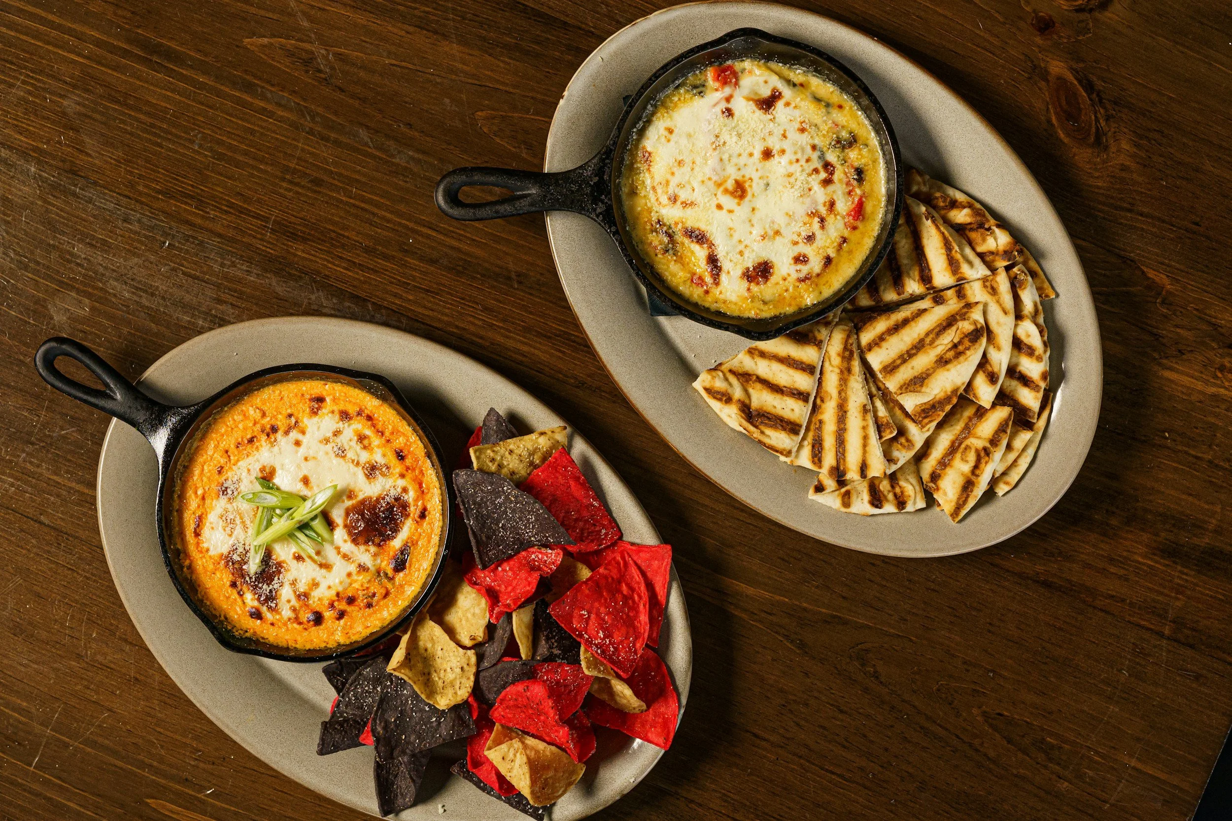 Two plates of baked cheese dip with grilled tortilla chips and colorful vegetable chips, set on a wooden table.