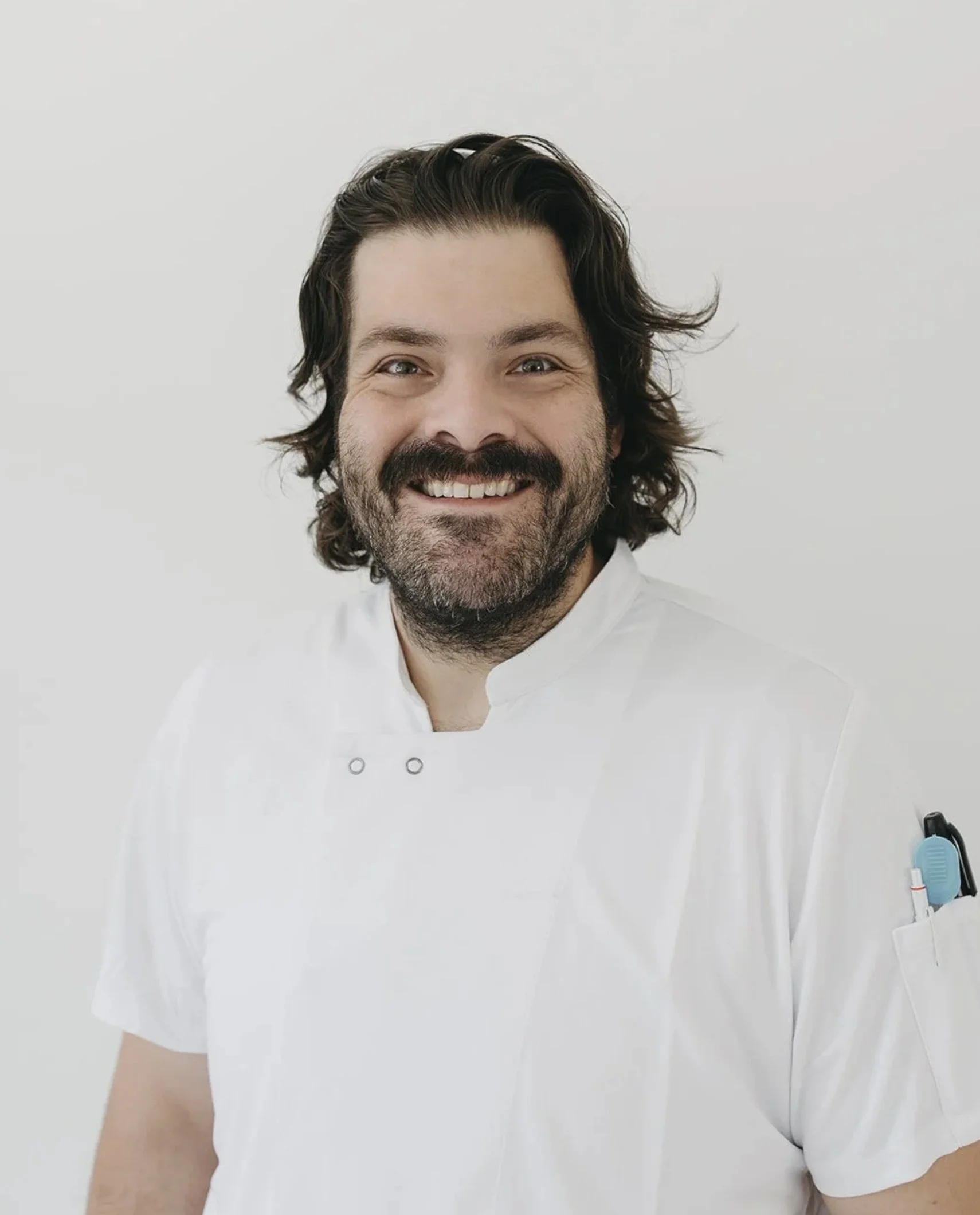 A man with long dark hair and a beard, dressed in a white shirt, smiling in front of a plain white background.