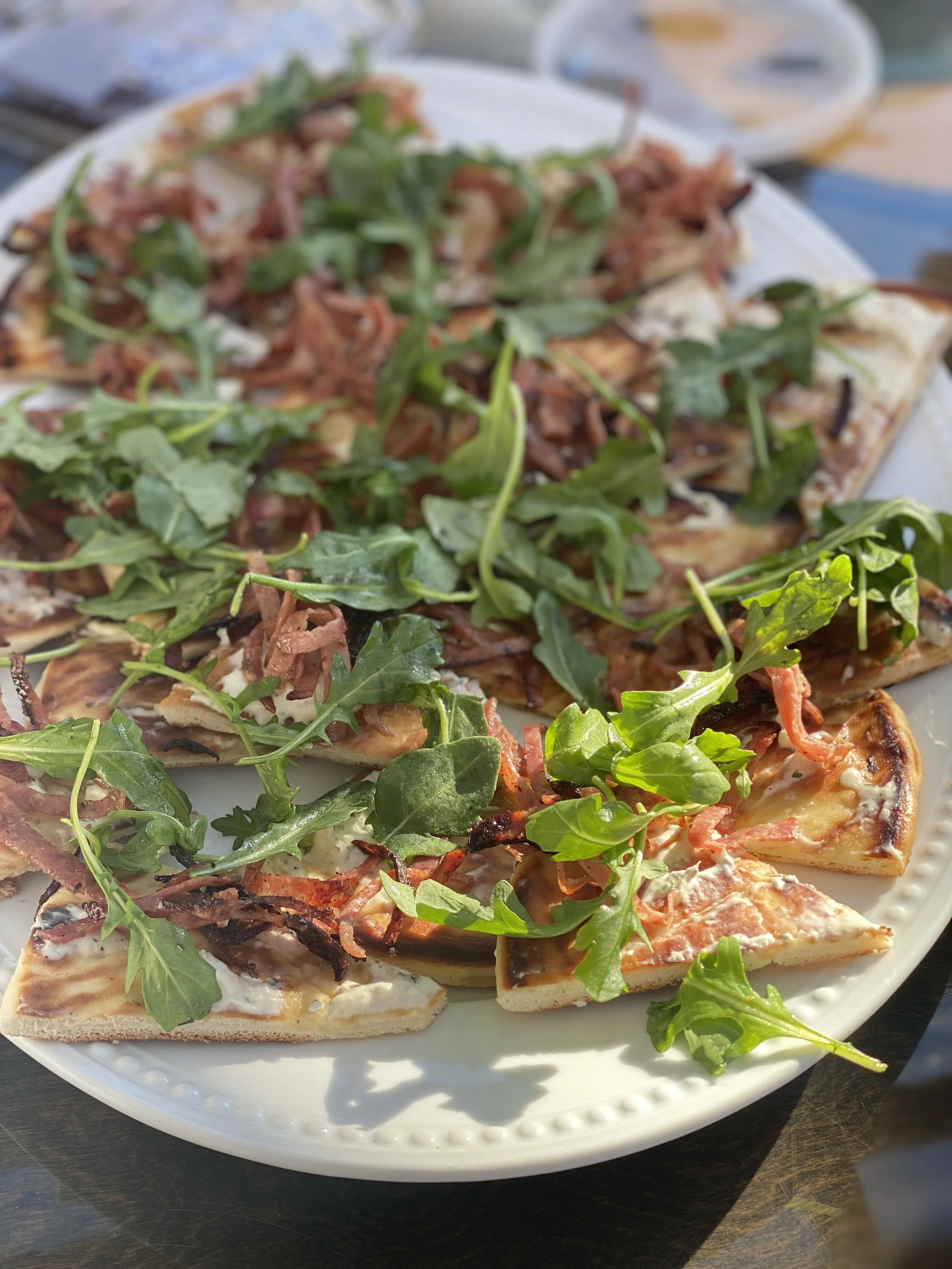 A white plate with slices of pizza topped with arugula, bacon, and cheese, placed on a dark surface with a bowl of sauce in the background.