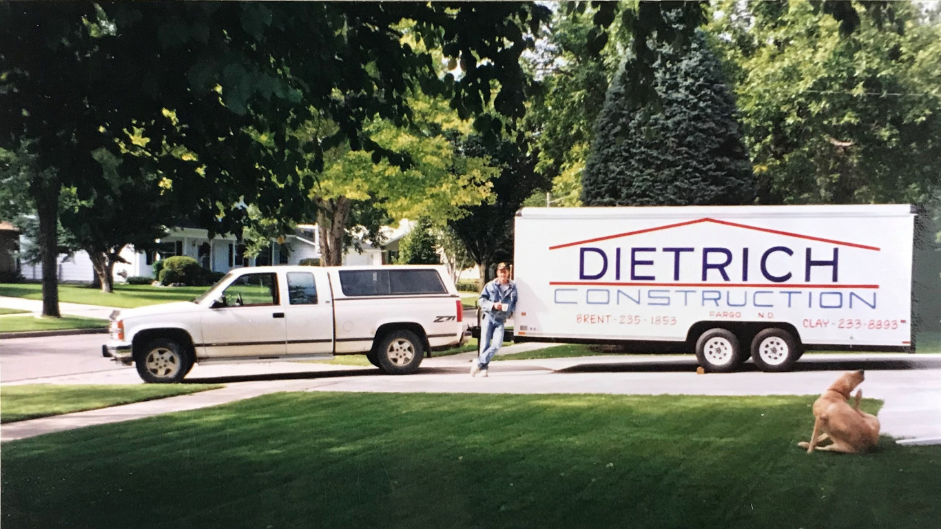 A man standing on the sidewalk next to a white pickup truck with a matching logoed trailer that reads 'Dietrich Construction.' A dog is lying on the grass nearby, and trees and houses are visible in the background.