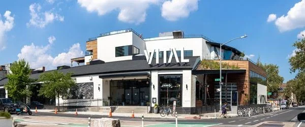 Modern building with a large sign that reads 'VITAL' on the front, situated on a street corner with trees and a clear sky in the background.