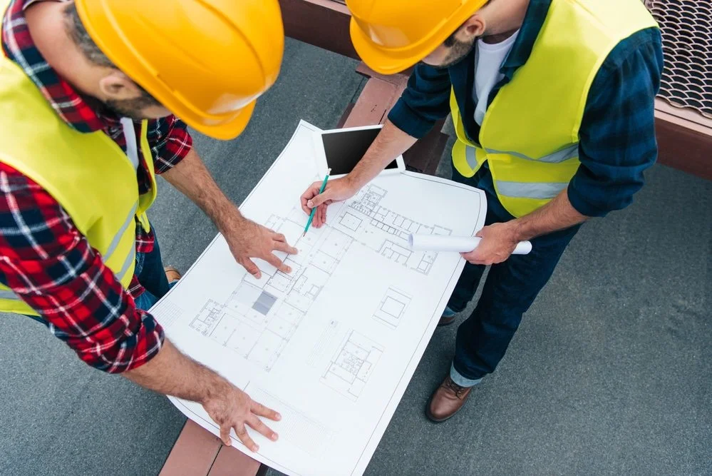 Two construction workers wearing yellow safety helmets and vests reviewing architectural blueprints on a table outdoors.