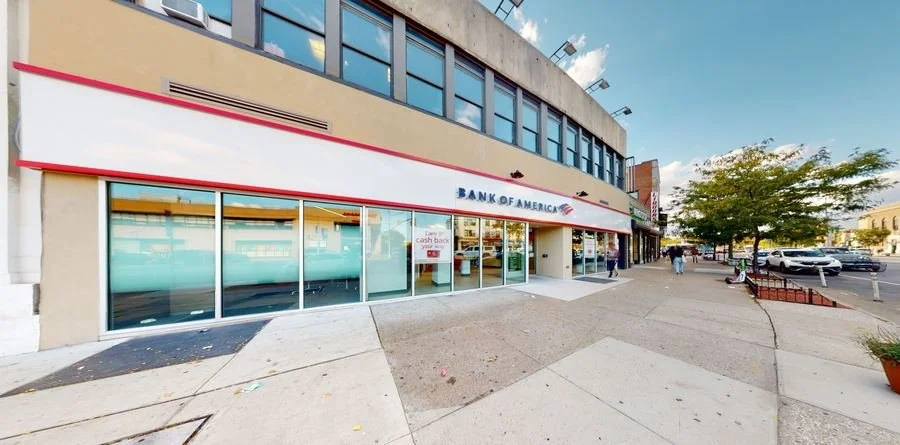 Exterior of a Bank of America branch with glass windows, sidewalk, and street view with cars and people.