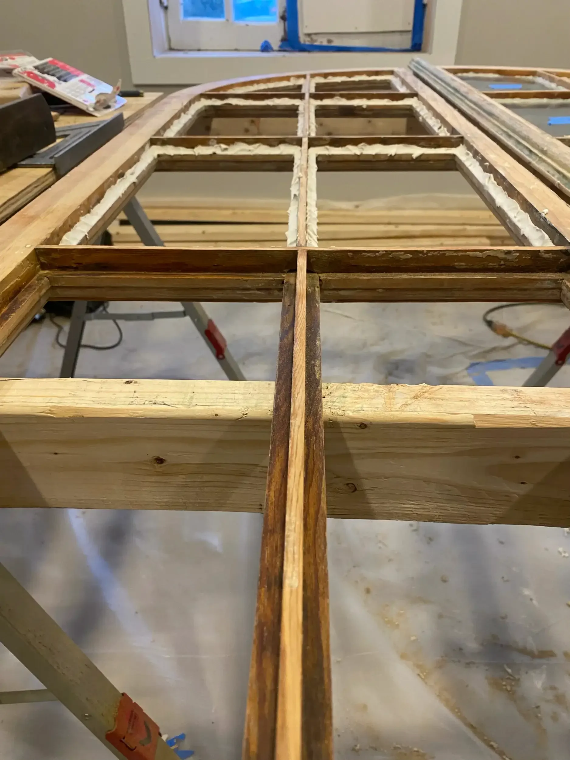 Interior view of a construction site showing a wooden window framework with foam insulation in the gaps, placed on work tables with construction tools, in a room with a window in the background.